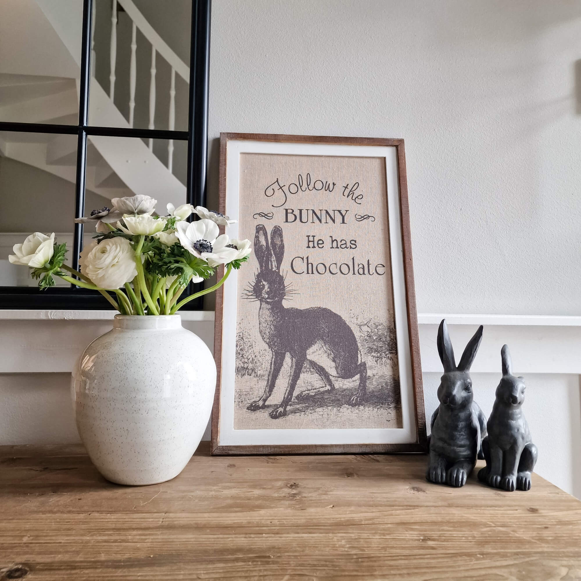 Decoratief schilderij met konijn en tekst, witte bloemen in keramische vaas, twee konijnenbeeldjes op houten tafel