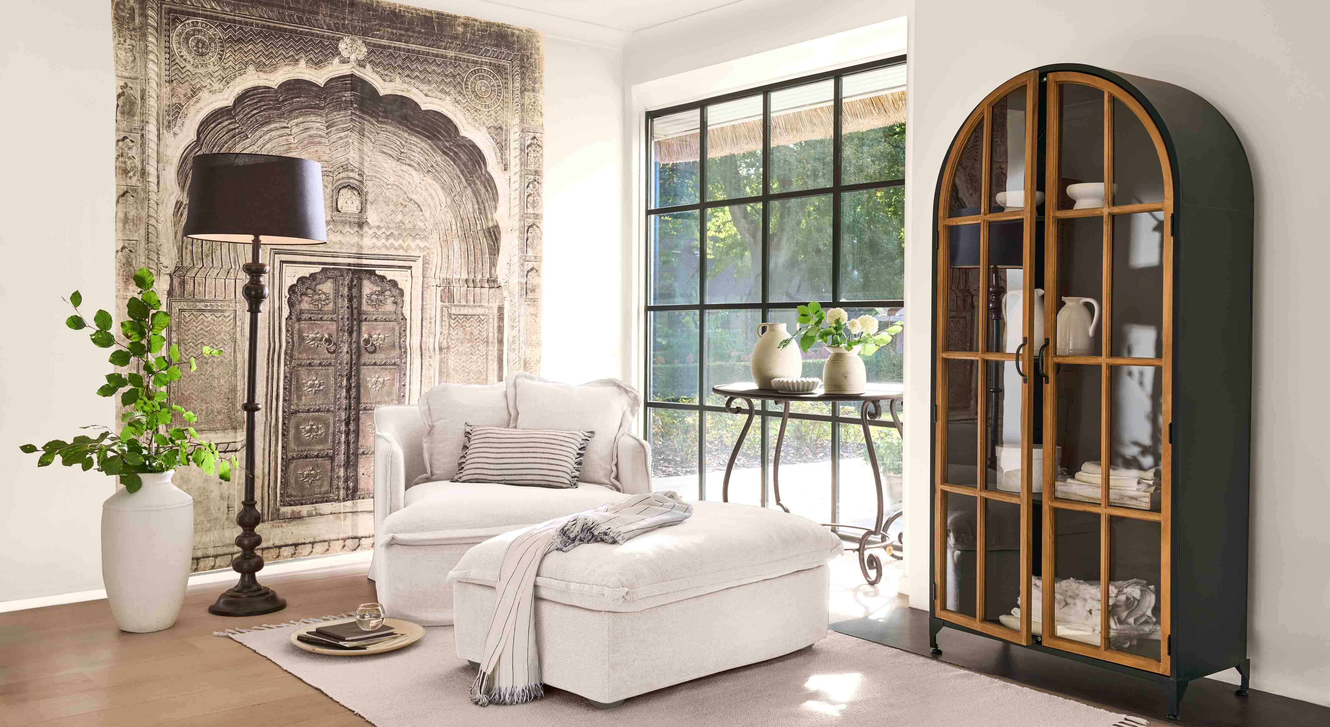 Comfortabele witte fauteuil met voetenbank en zwarte staande lamp in lichte woonkamer met decoratieve kast