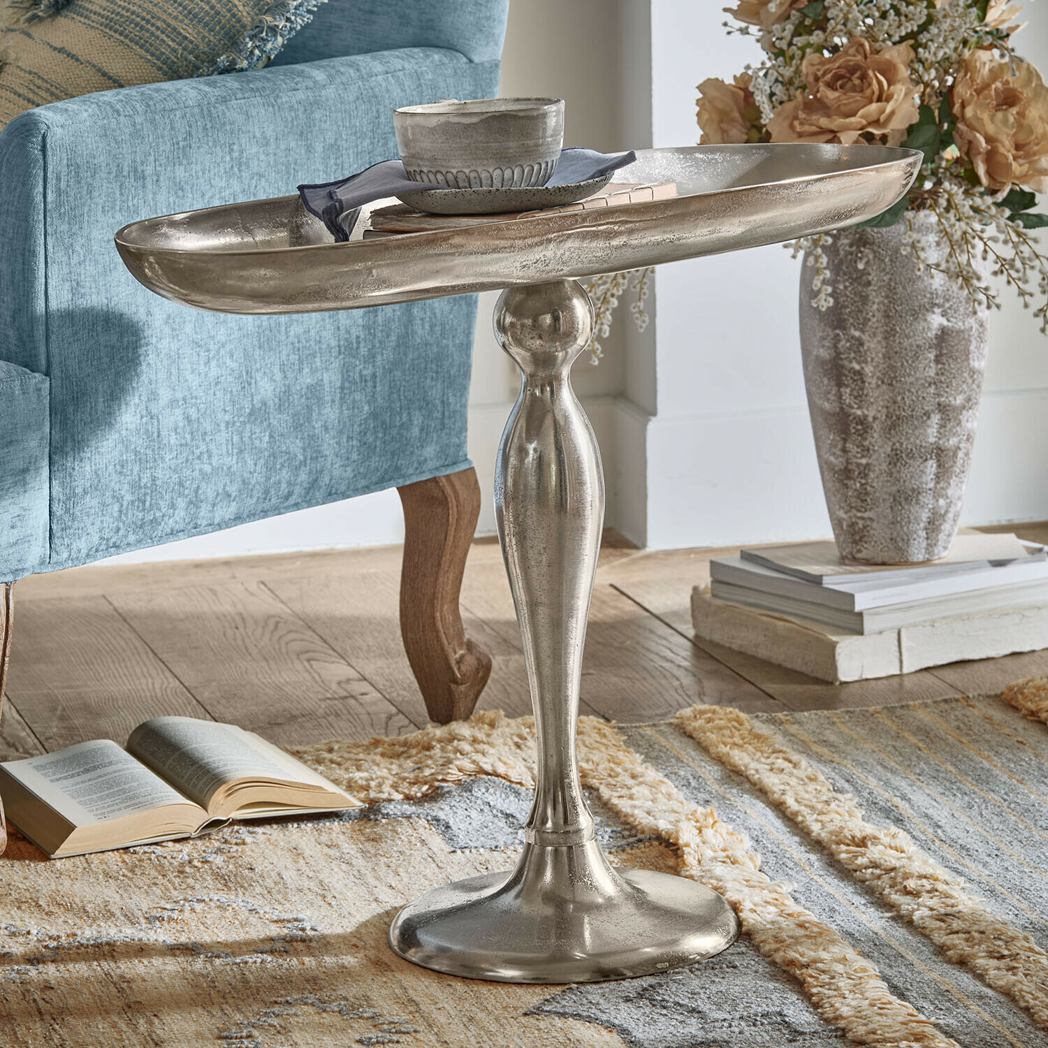 Zilverkleurige metalen bijzettafel met decoratieve voet op tapijt naast blauwe fauteuil