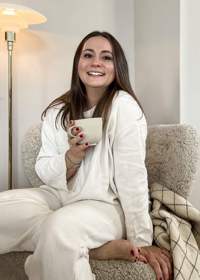 Lachende vrouw in witte zachte vrijetijdskleding met mok zittend op een comfortabele stoel in een warme kamer