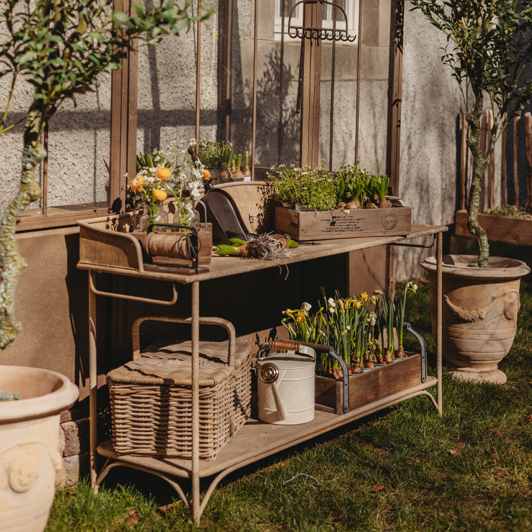 Metalen tuintafel met houten kisten en bloempotten op gras bij een kasmuur