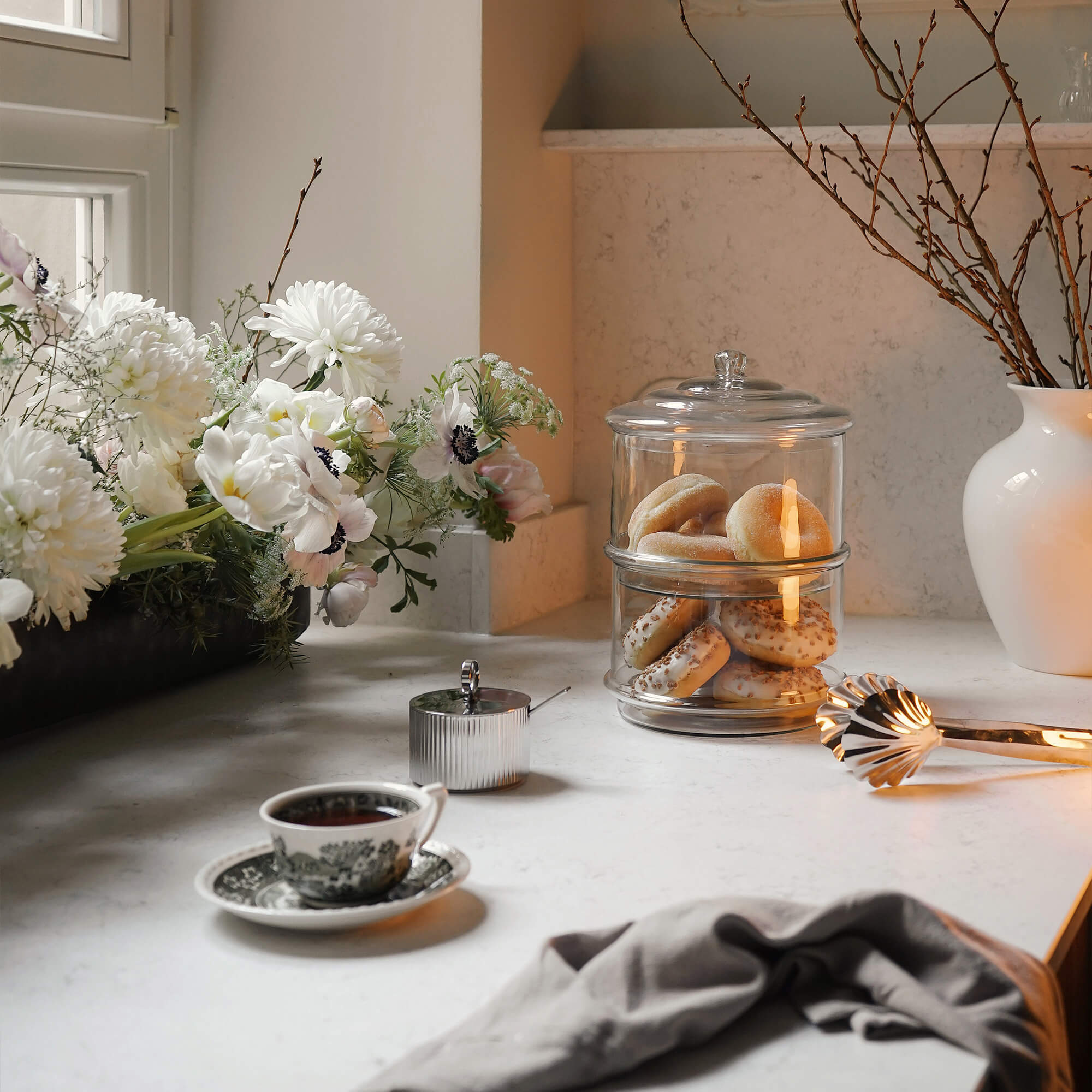 Glazen koekjespot met twee lagen op een lichte keukenblad met bloemen en theekopje
