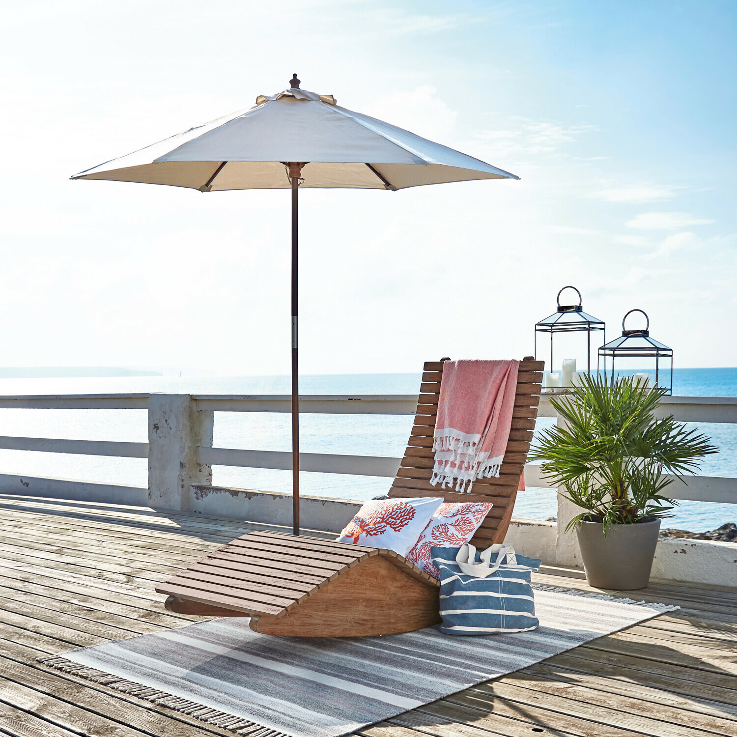 Parasol Enfield Houten ligstoel met witte parasol en decoratieve kussens op terras aan zee, perfect voor zomerse ontspanning