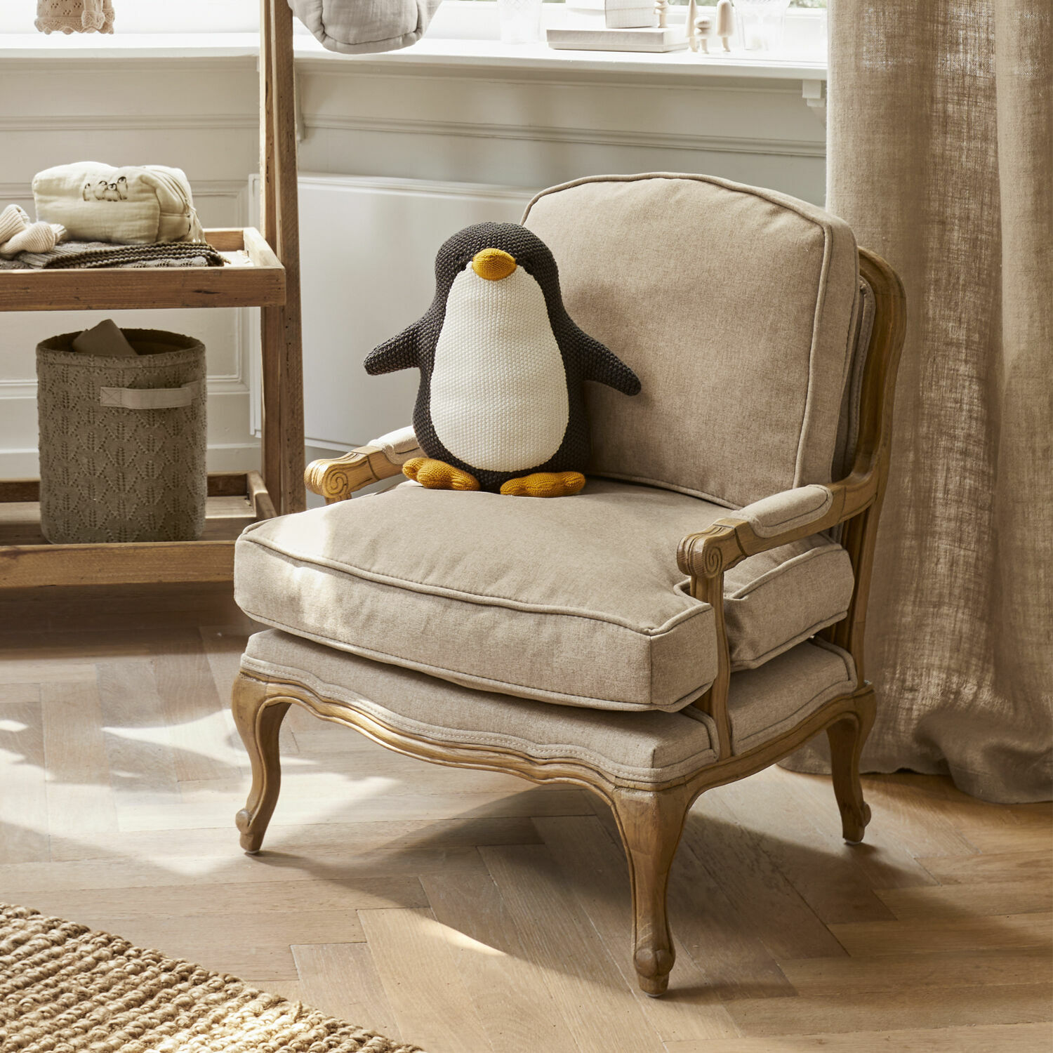Beige fauteuil met houten frame en zacht kussen met een gebreide pinguïn knuffel