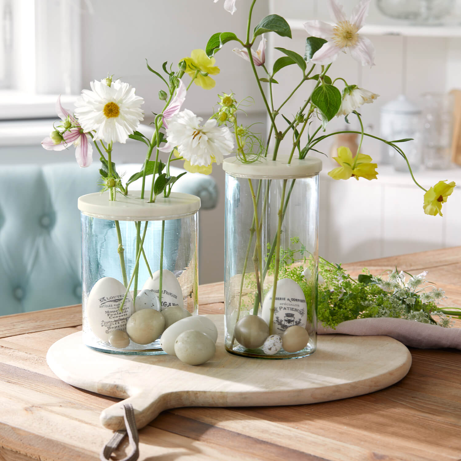 Glazen vazen met crèmekleurige deksels en decoratieve eieren, pastelkleurige bloemen op een lichte houten plank