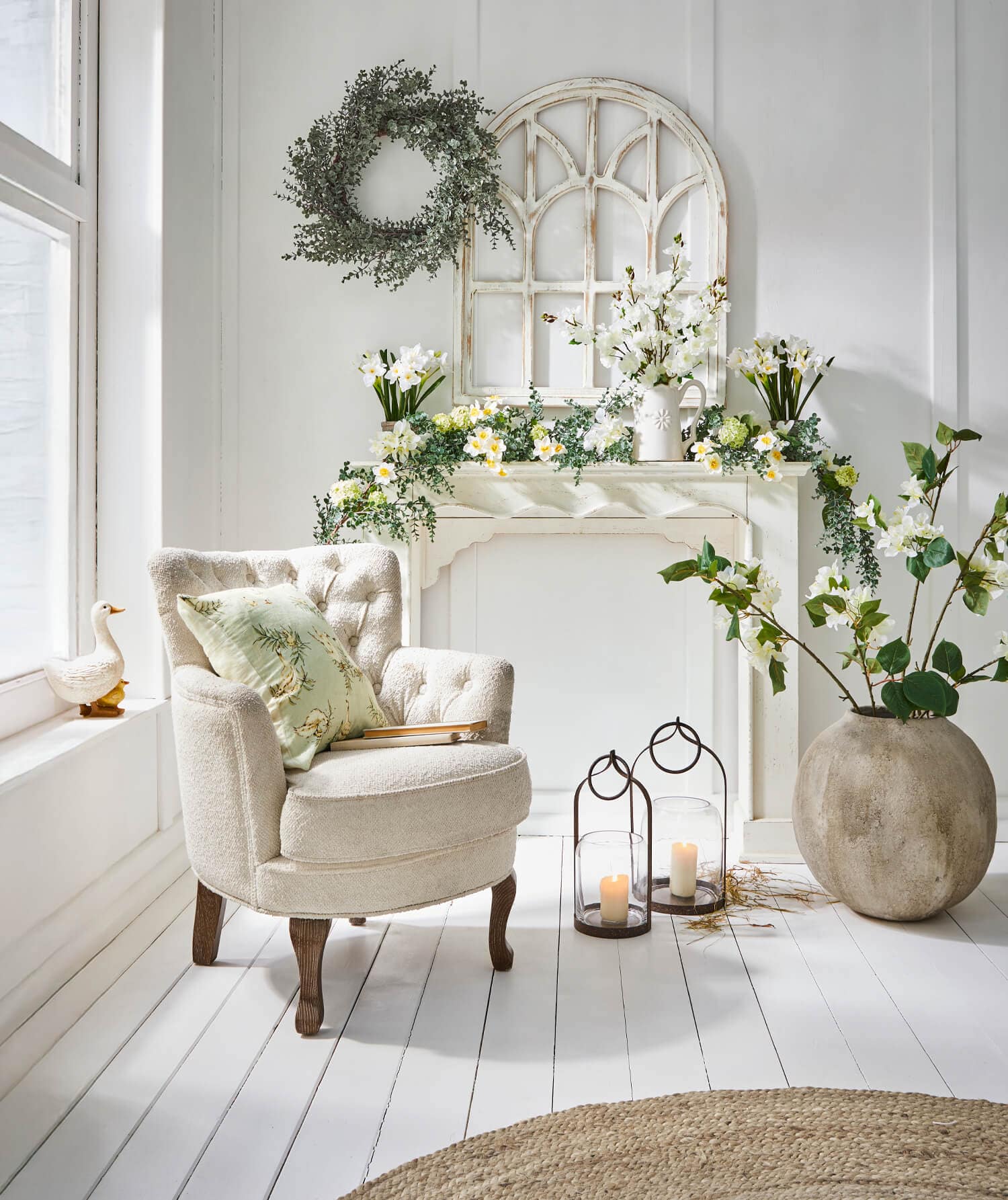 Beige stoffen fauteuil met kussen naast decoratieve schoorsteenmantel met bloemen en lantaarns in lichte kamer