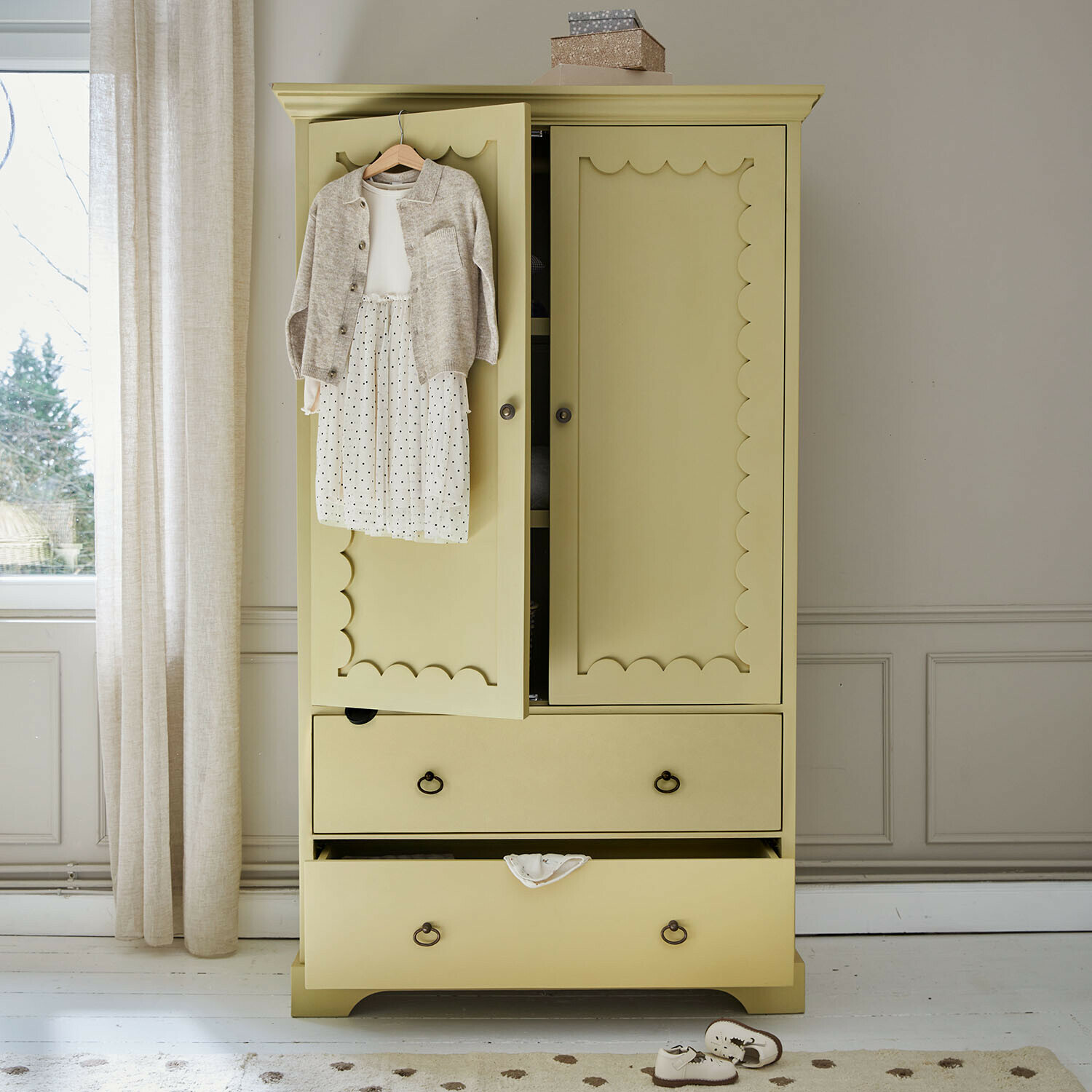 Kledingkast voor kinderen Frollin Gele houten kast met decoratieve deuren en kinderkleding aan de deur in een lichte kamer