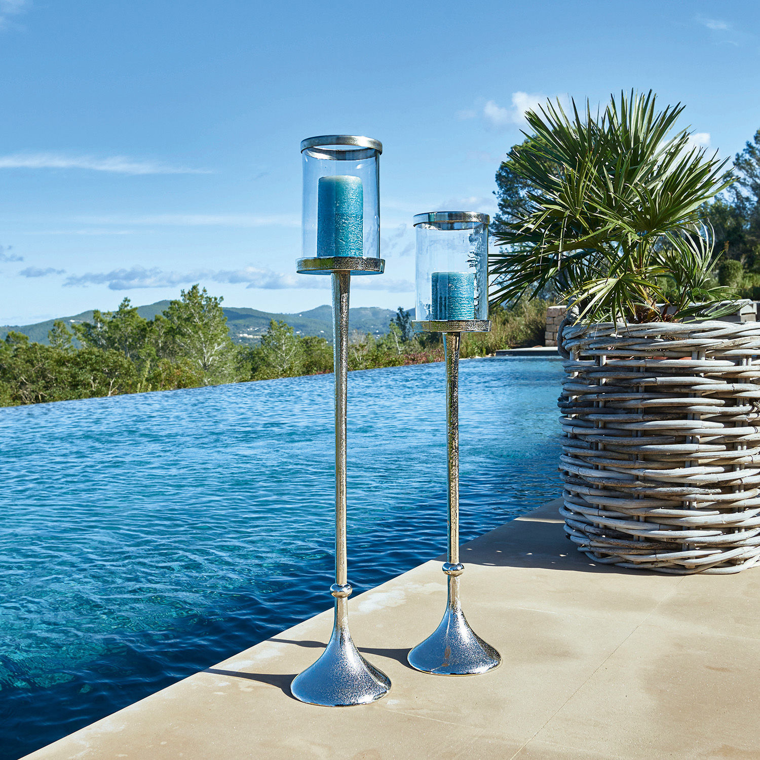 Twee zilverkleurige metalen kandelaars met blauwe kaarsen bij het zwembad naast een plant in een rieten mand