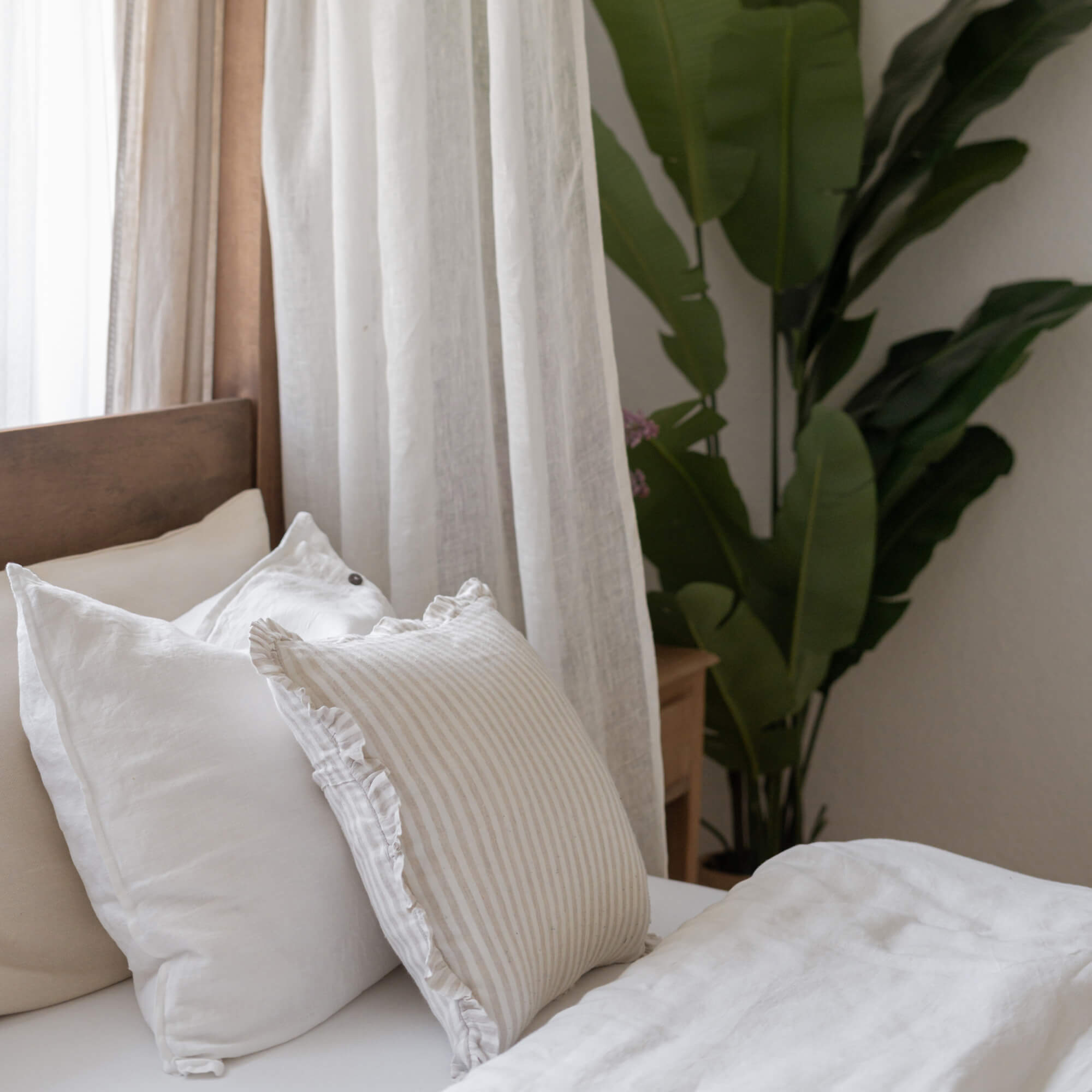 Gezellige witte en beige linnen kussens op bed met houten hoofdeinde en groene plant op de achtergrond