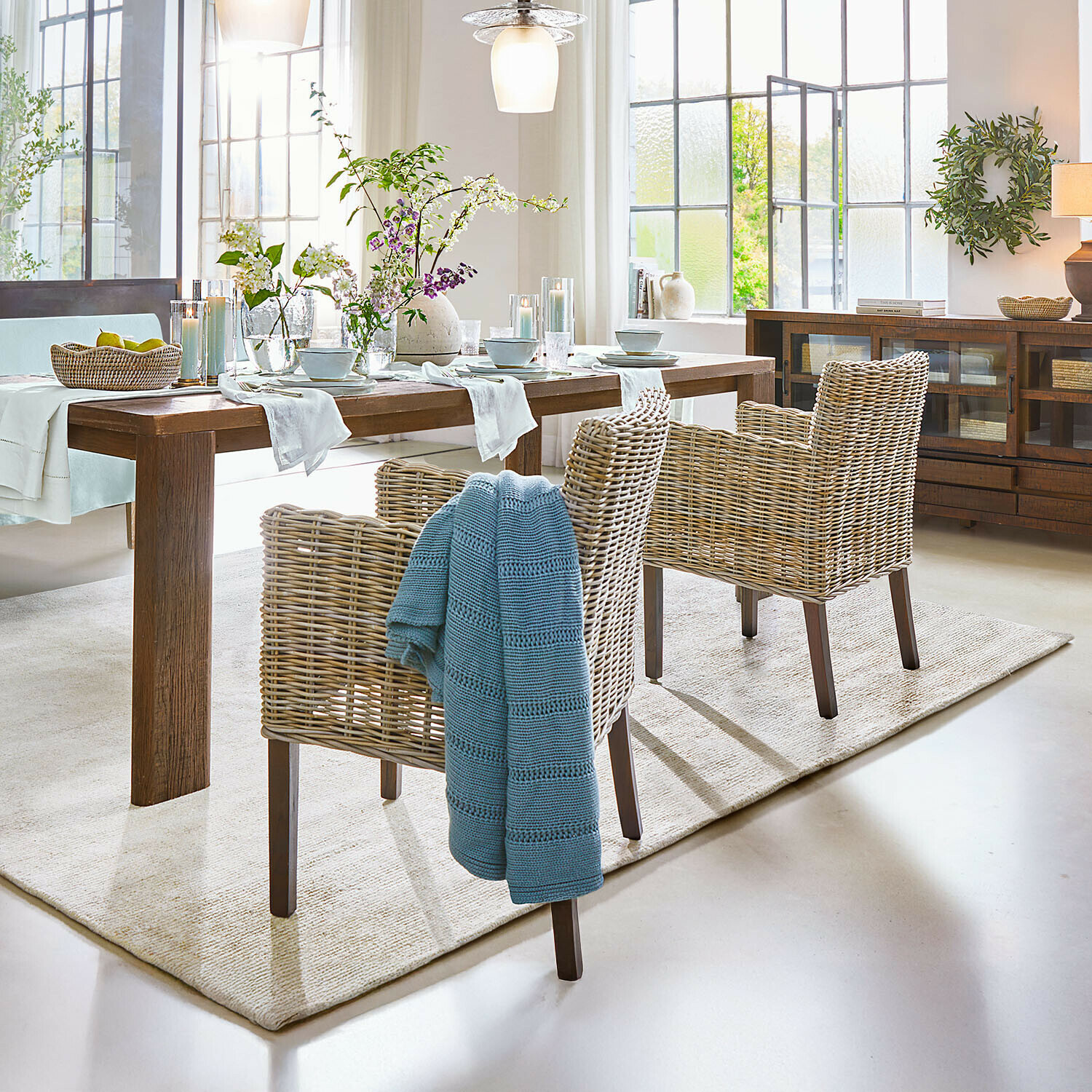 Eetkamer met houten tafel en natuurlijke rotan stoelen, bloemen en blauwe deken