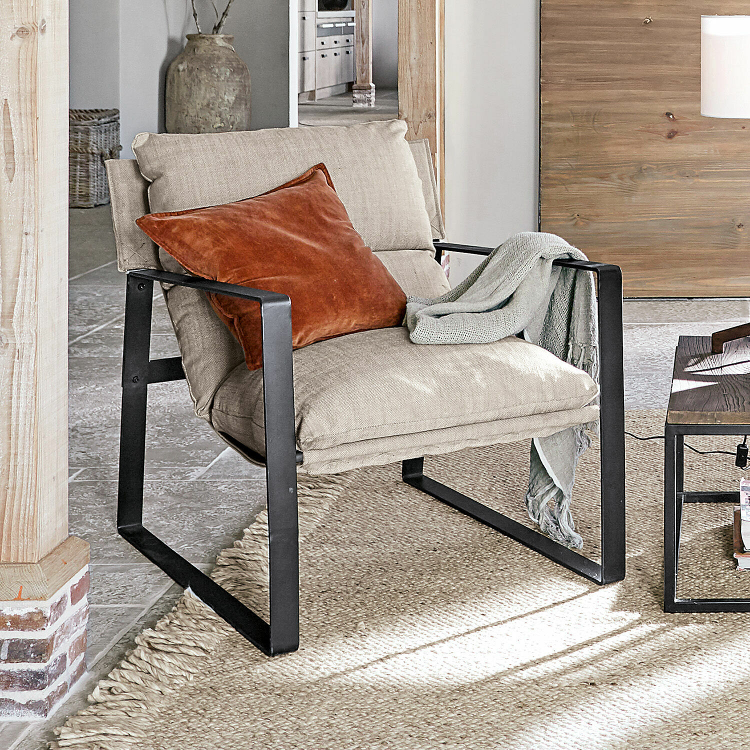 Fauteuil Seuvard Beige stoffen fauteuil met zwart metalen frame, oranje kussen en grijze deken in woonkamer