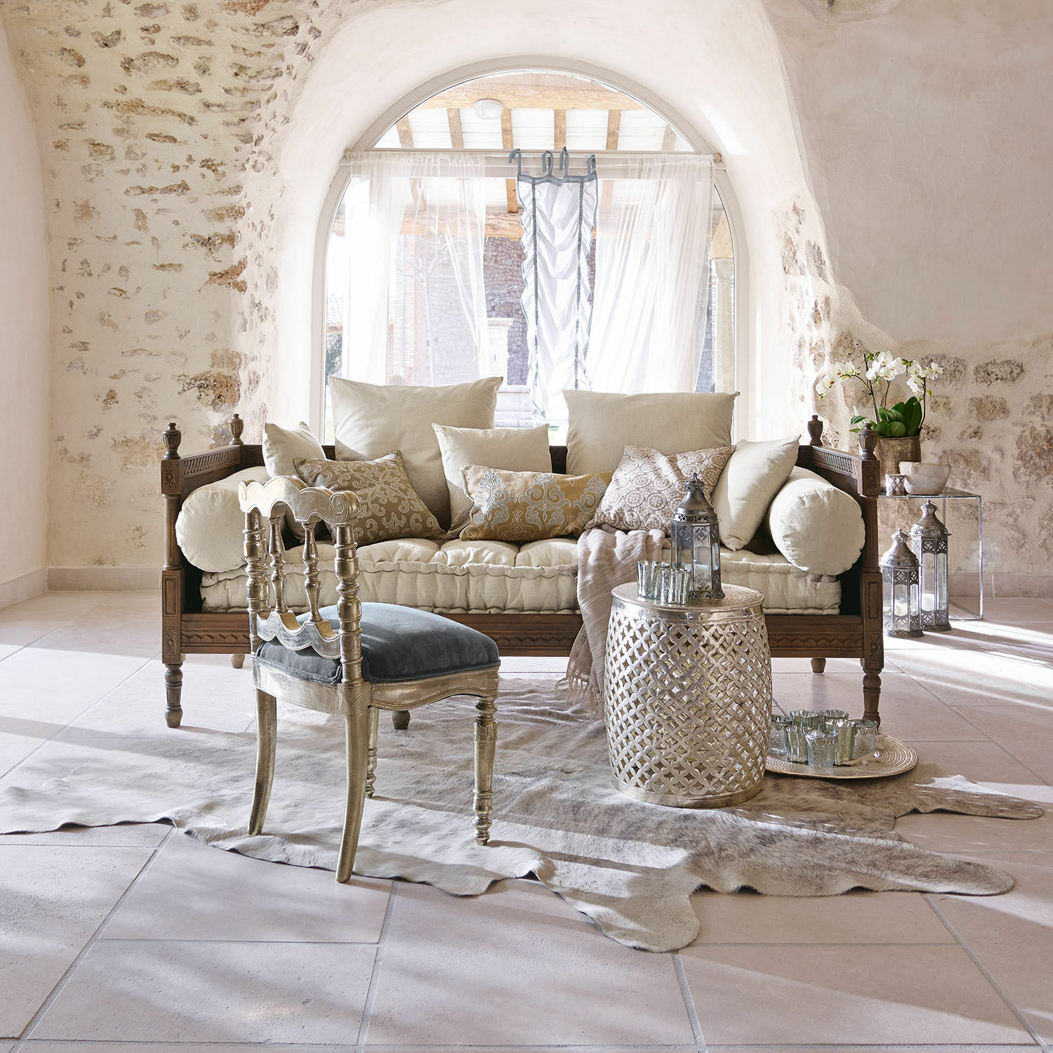 Gezellige woonkamer met houten bank, crèmekleurige kussens, zilveren bijzettafel en blauwe fluwelen stoel