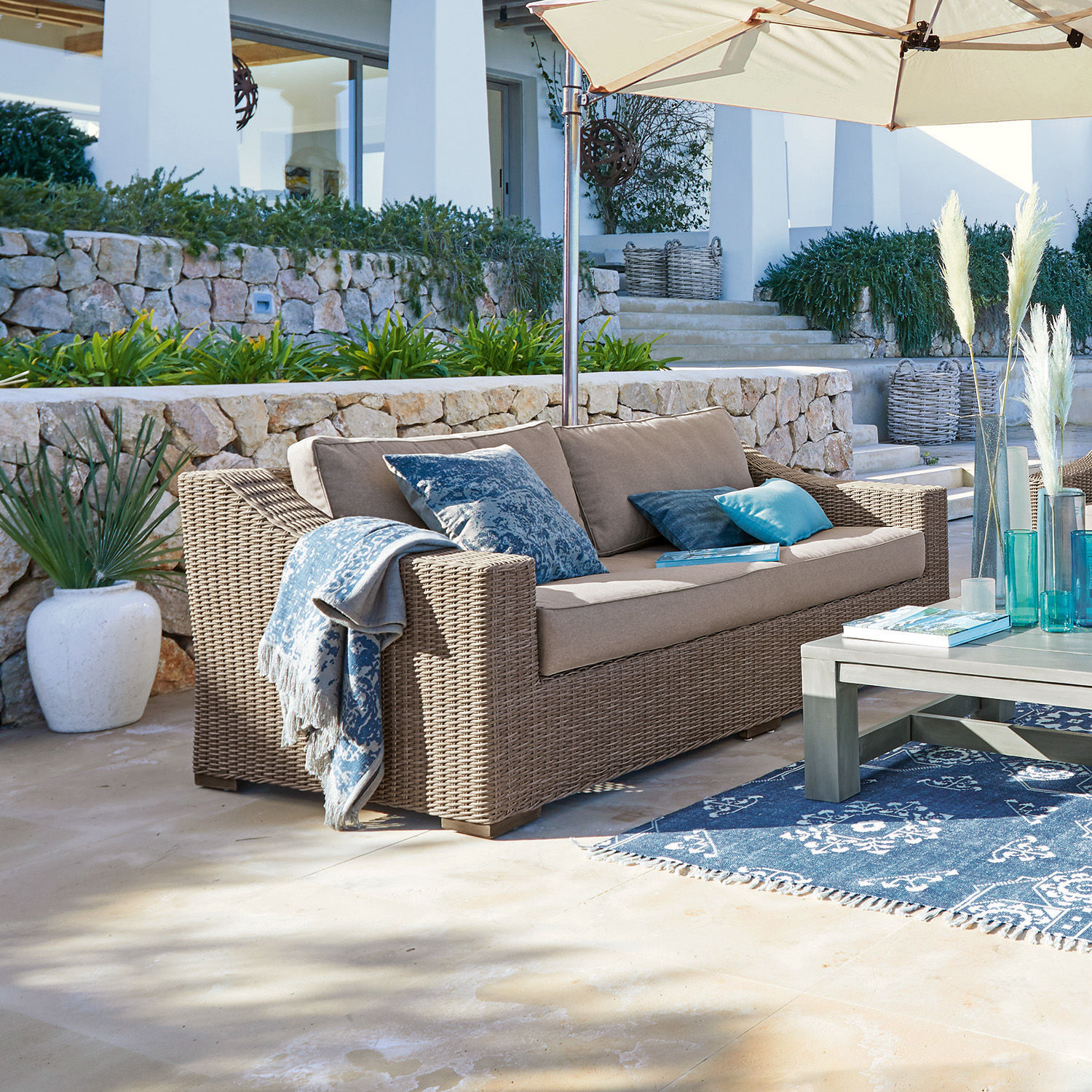 Bank Chestingdale Beige rattan loungesofa met blauwe kussens en parasol op zonnig terras