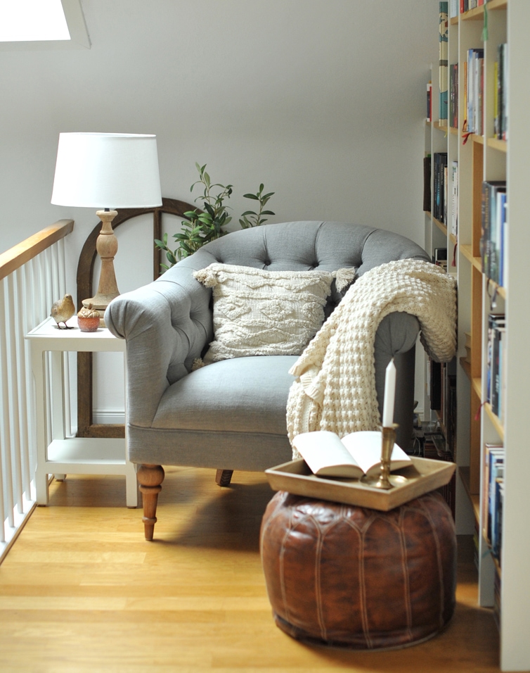 Grijze gestoffeerde fauteuil met crème kussen en deken, bruine leren poef en lamp op wit tafeltje