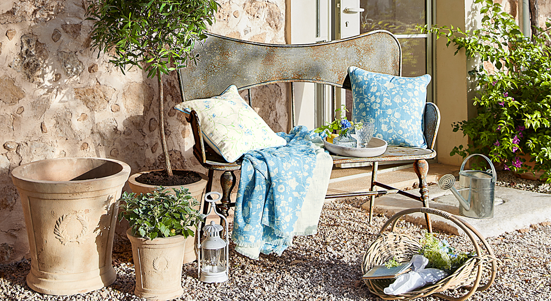 Rustieke metalen bank met blauwe kussens en deken in Provençaalse tuin met potten en gieter