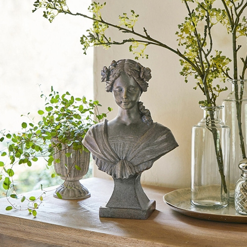 Grijs stenen vrouwenbuste met bloemen in het haar op houten tafel met planten decoratie