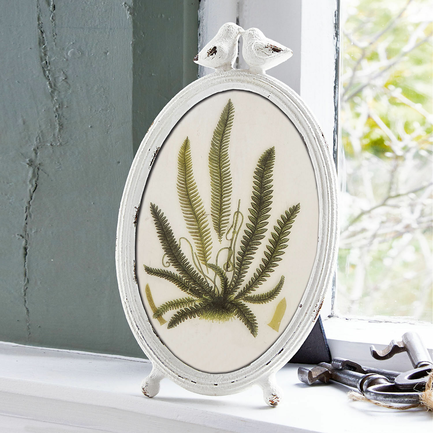 Ovale witte metalen lijst met vogeldecoratie en groen varenmotief op vensterbank