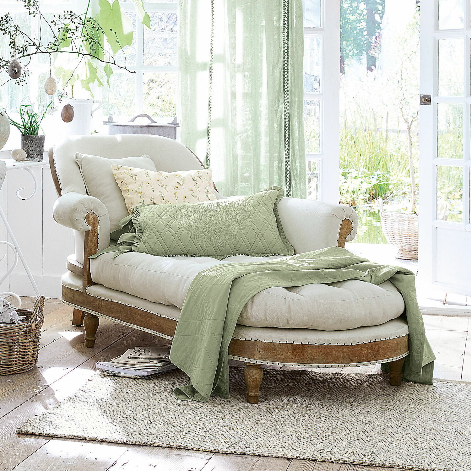 Elegante chaise longue van licht hout en crème stof met groene deken en kussens in lichte kamer