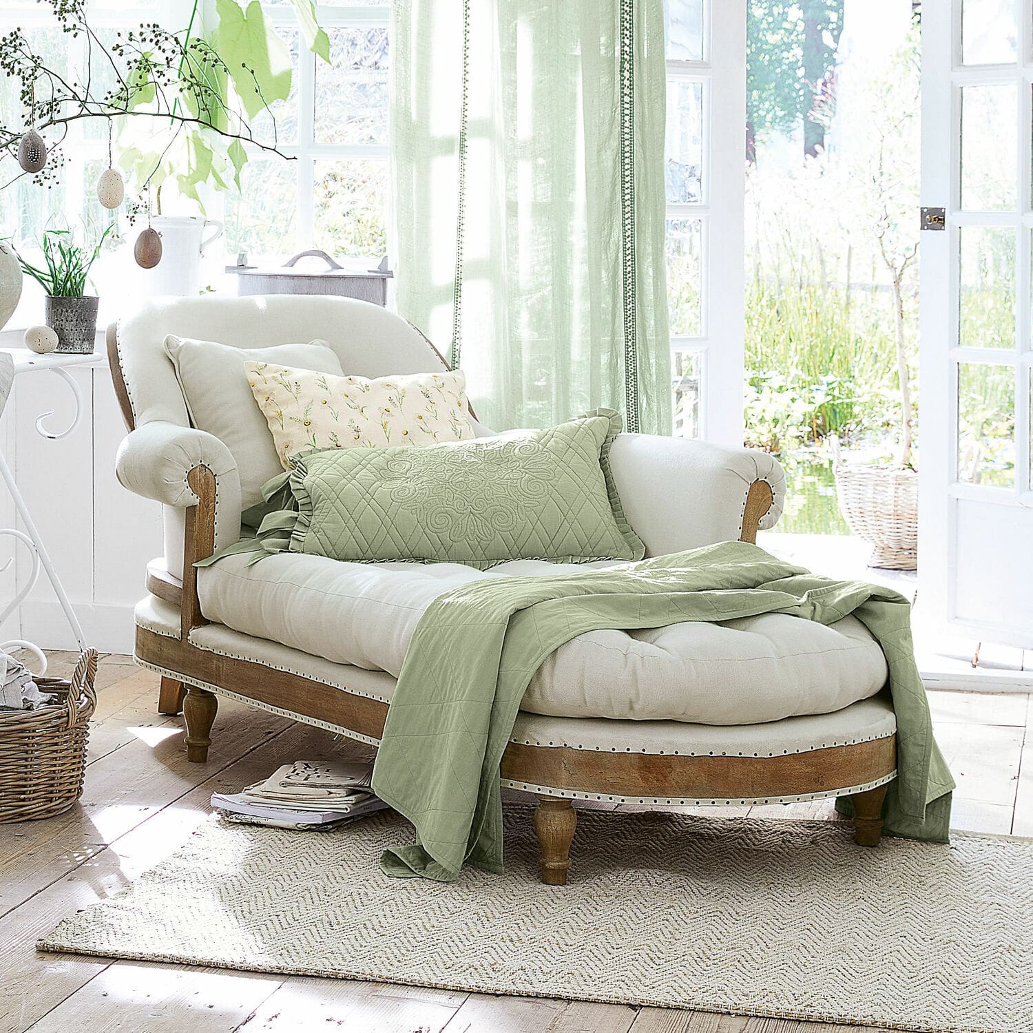 Chaise longue Queyranne Elegante chaise longue van licht hout en crème stof met groene deken en kussens in lichte kamer