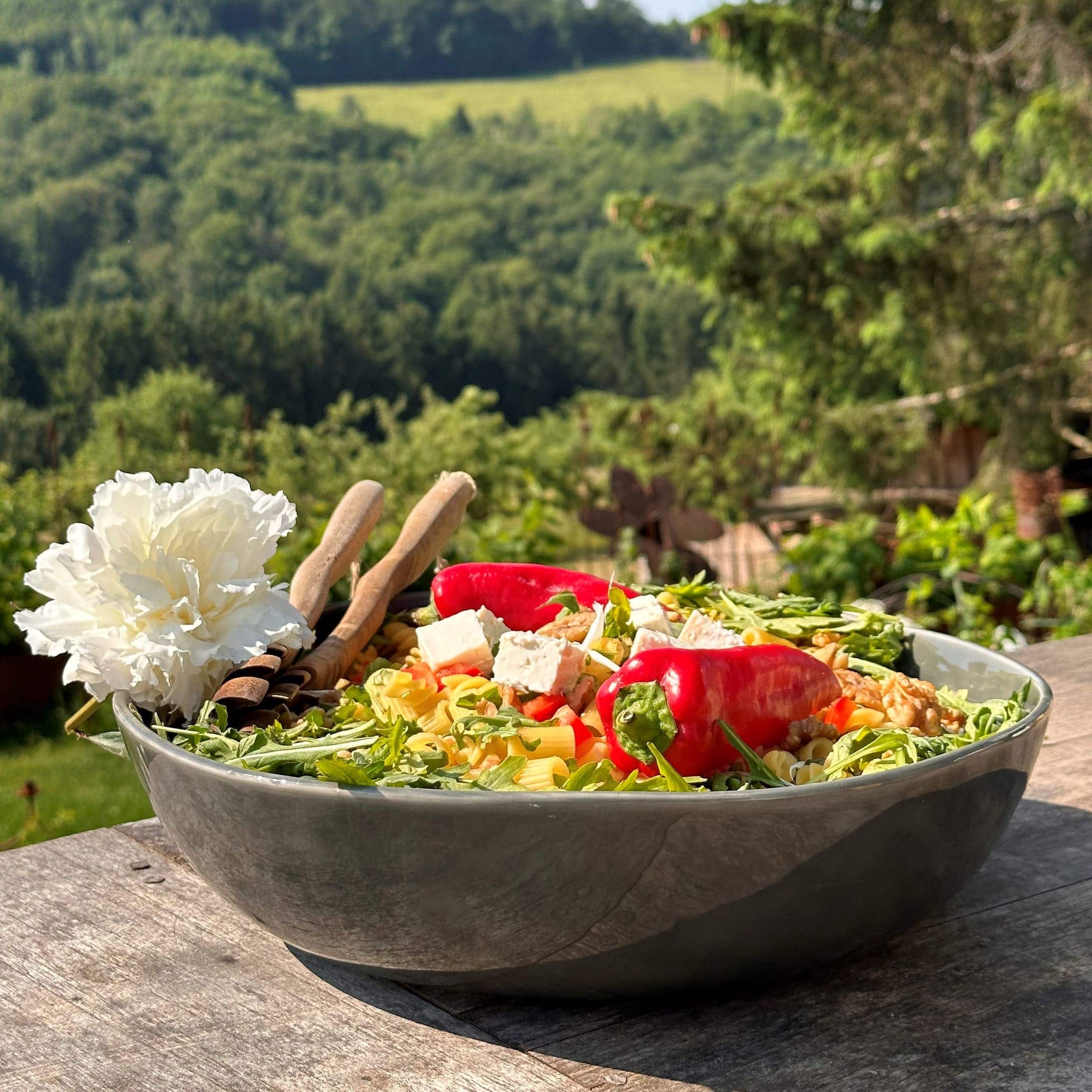 Grote keramische kom met salade, rode paprika, feta en rucola op houten tafel buiten