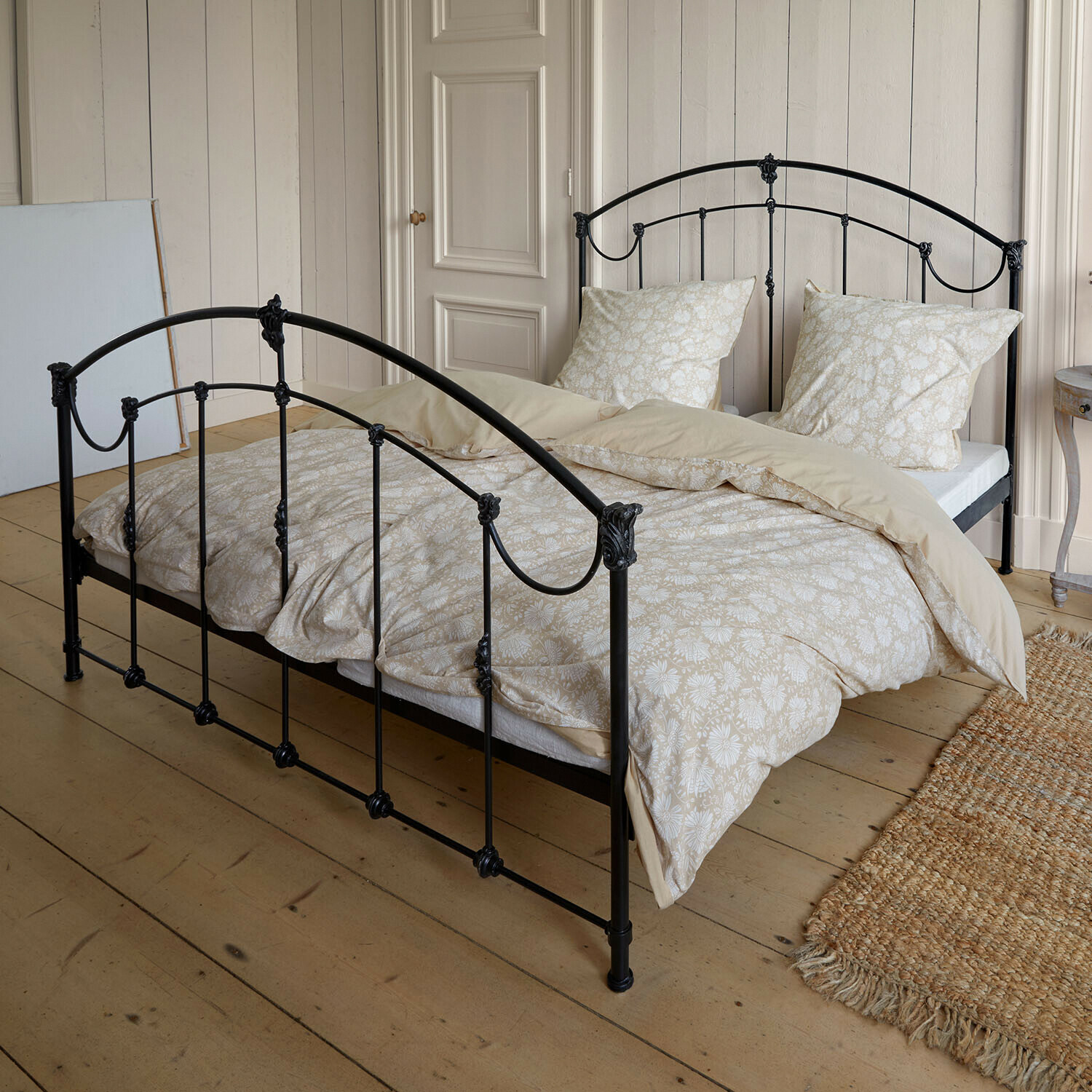 Bed Canefield Zwart metalen bed met beige beddengoed met bloemenpatroon op lichte houten vloer