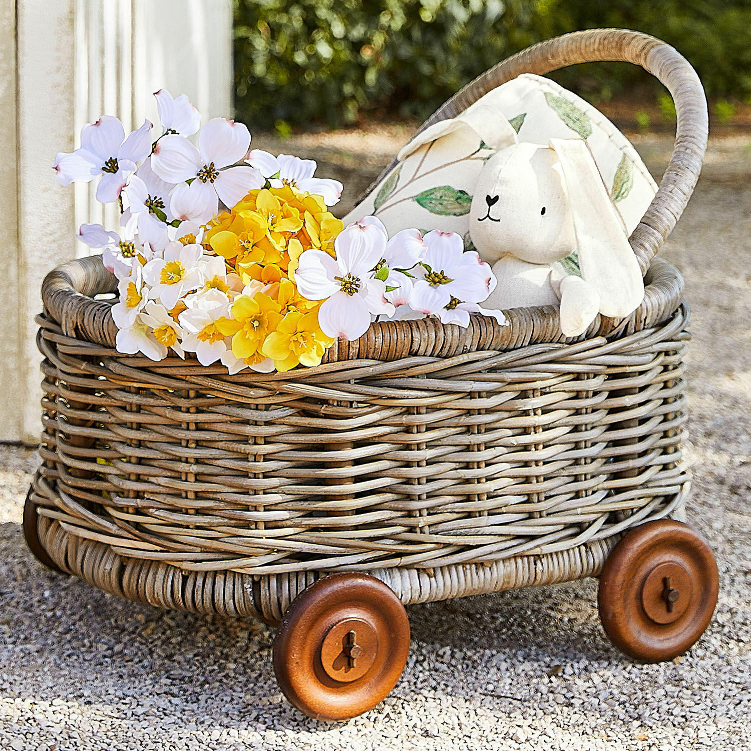 Trolley Narivena Rieten karretje met houten wielen, gevuld met gele en witte bloemen en een beige knuffelkonijn