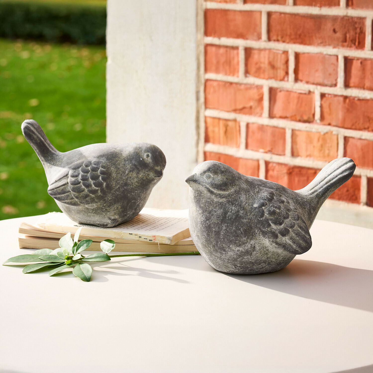 Twee grijze stenen vogelbeeldjes op een witte tafel met boek en groen blad als decoratie