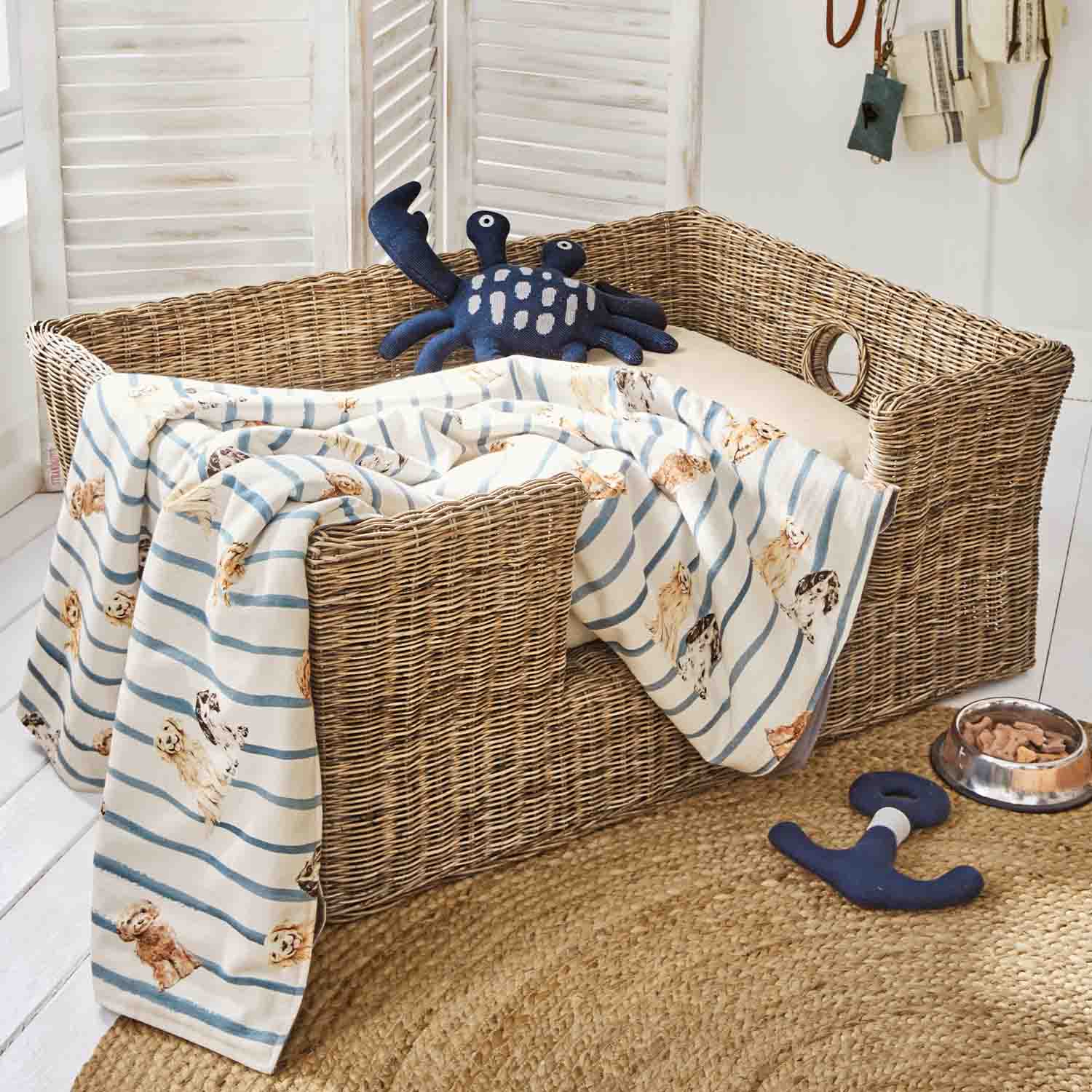 Rieten hondenmand met crème kussen, gestreept hondenprintdeken en donkerblauw krabvormig speeltje