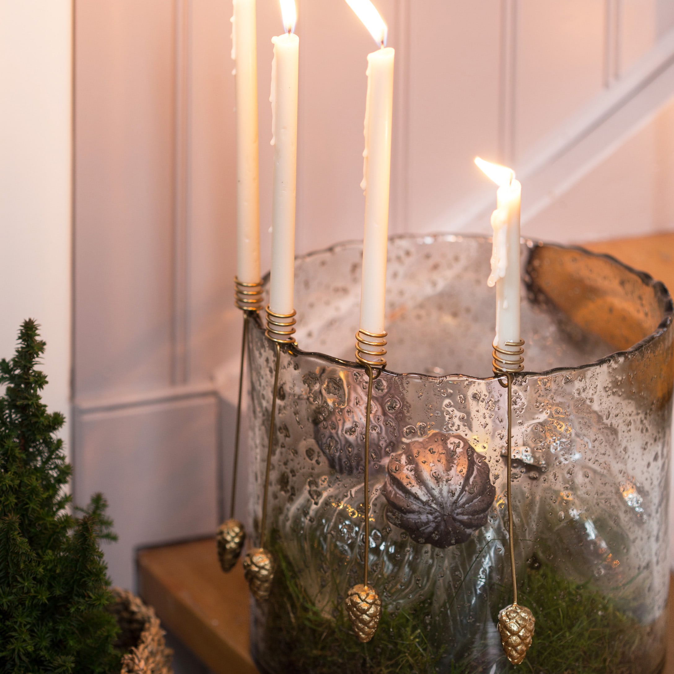 Glazen kandelaar met witte kaarsen en gouden dennenappelversieringen op groen mos