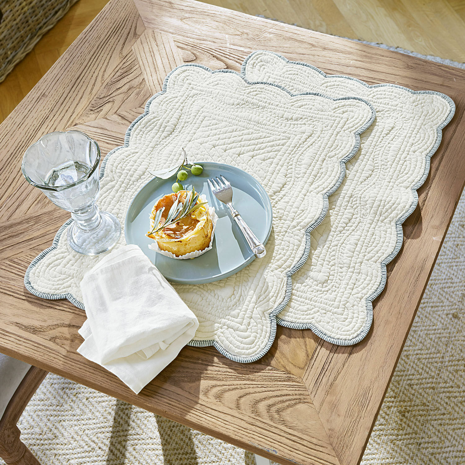 Placemat set van 2 Fontienne Beige gewatteerde katoenen placemats met blauwe rand op houten tafel met dessert en glas