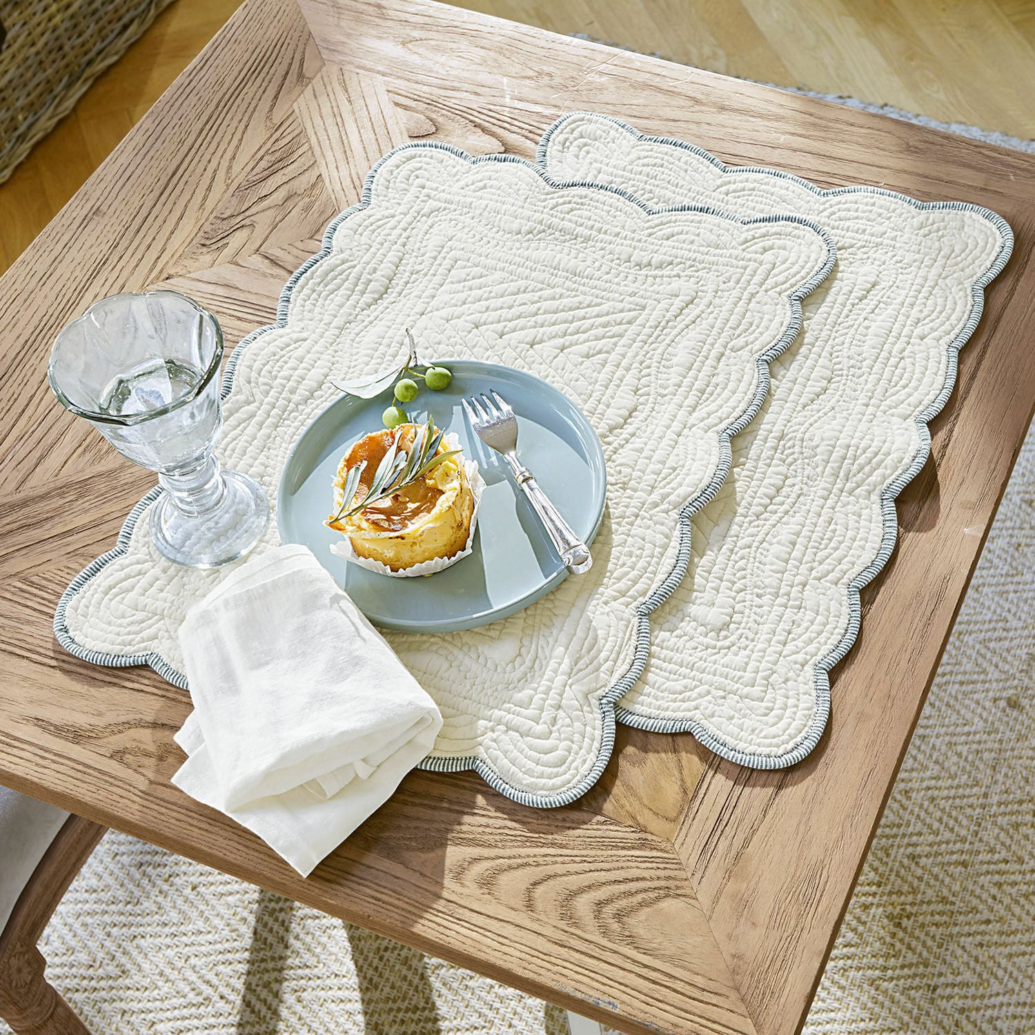 Placemat set van 2 Fontienne Beige gewatteerde katoenen placemats met blauwe rand op houten tafel met dessert en glas