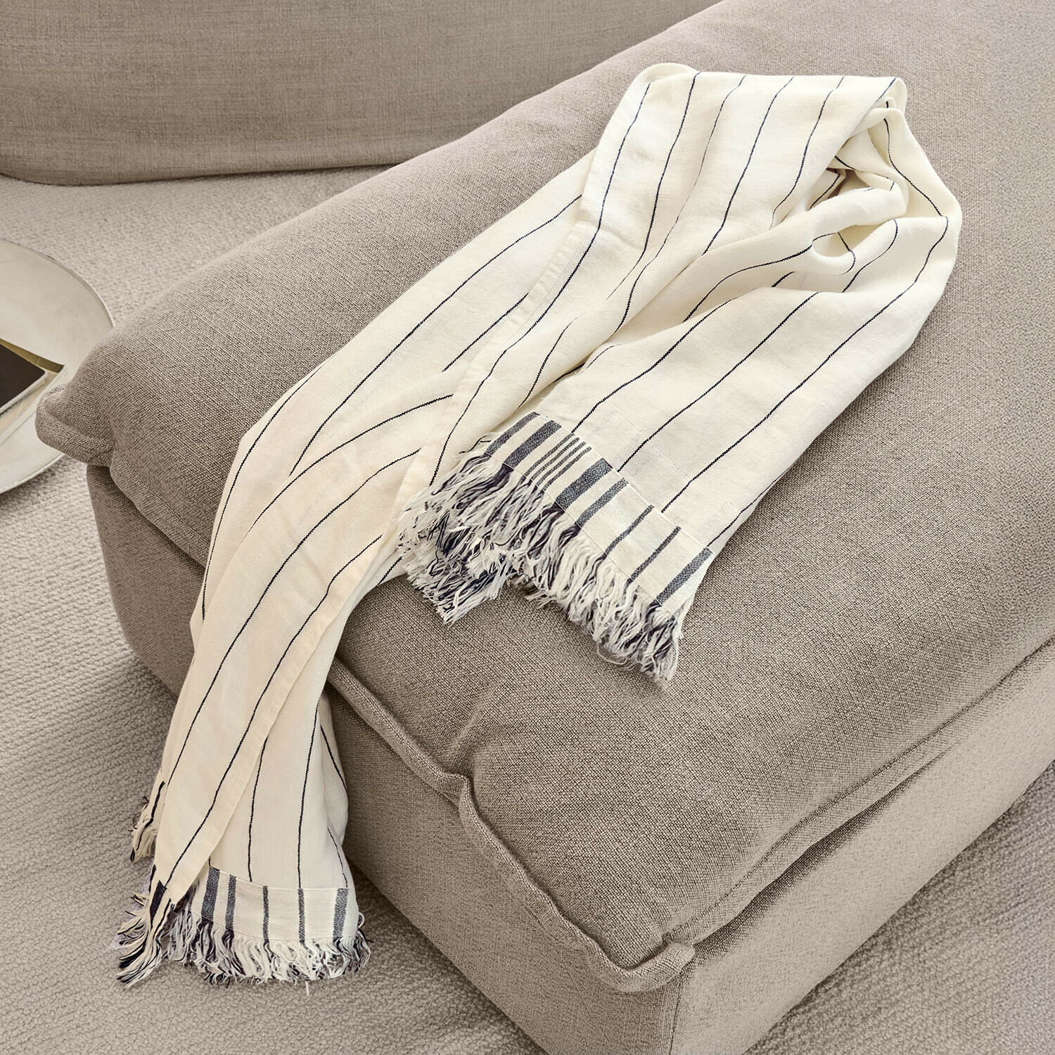 Plaid Ulderina Beige katoenen deken met zwarte strepen en franjes op een grijze stoel, sfeervolle woondecoratie