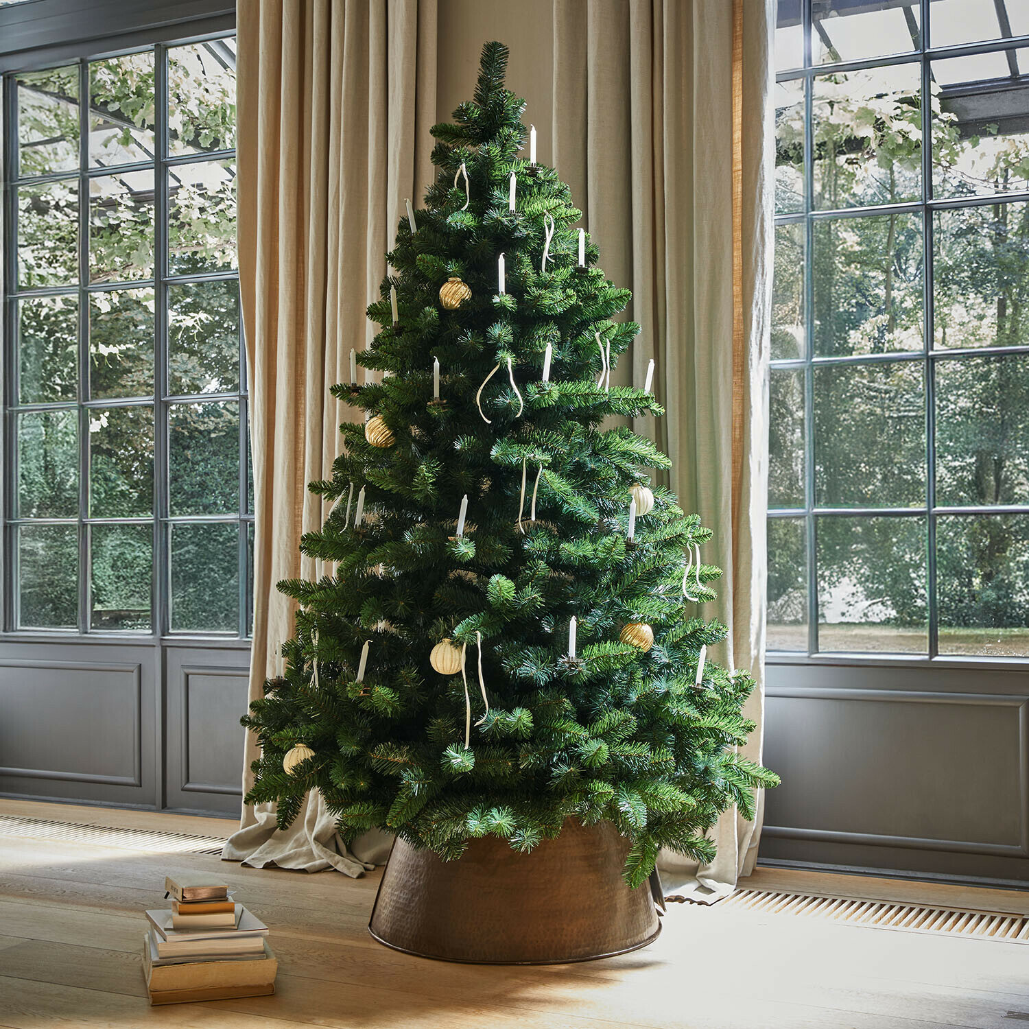 Kunstkerstboom met groene takken en witte kaarsen in koperen pot bij raam met beige gordijnen