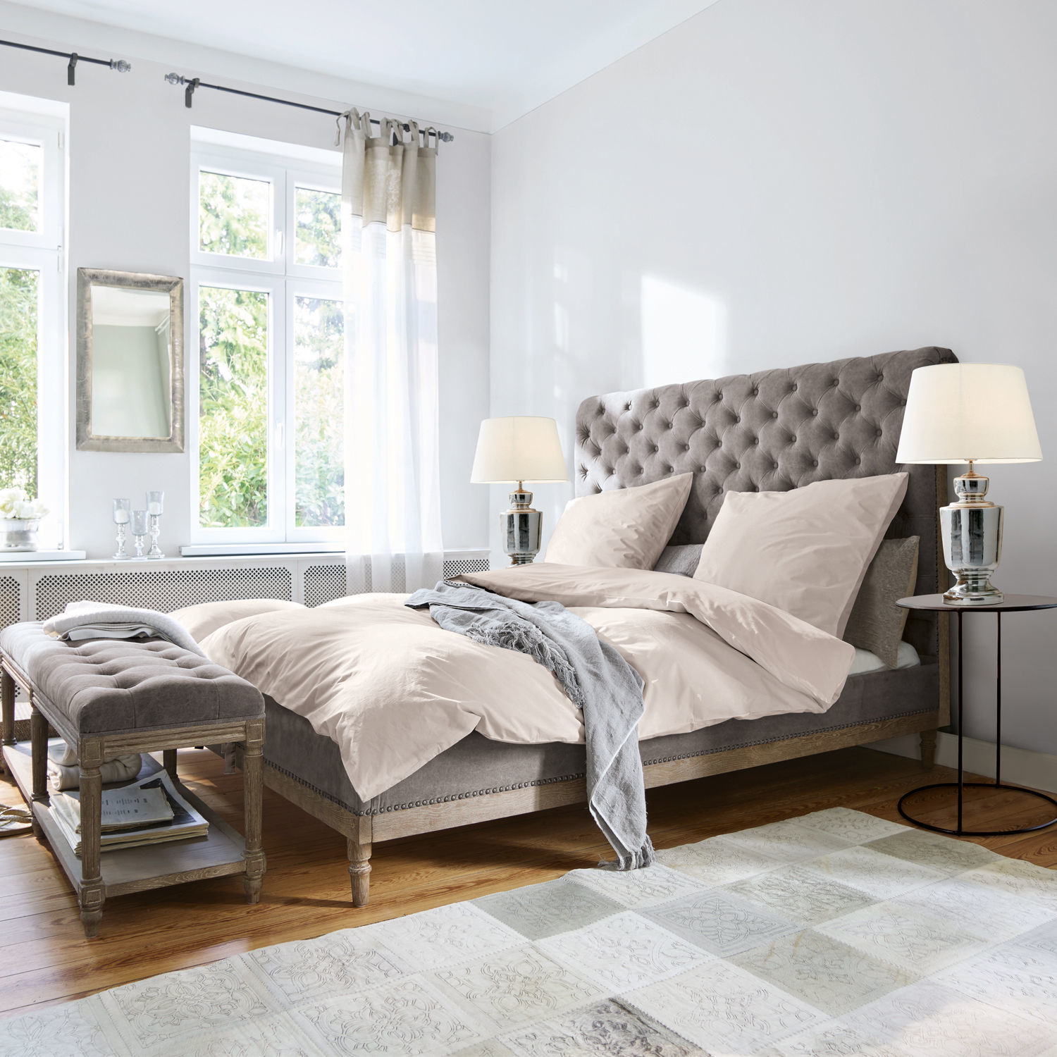Bed Pozières Grijs gestoffeerd bed met doorgestikt hoofdeinde en beige beddengoed in lichte slaapkamer met houten vloer