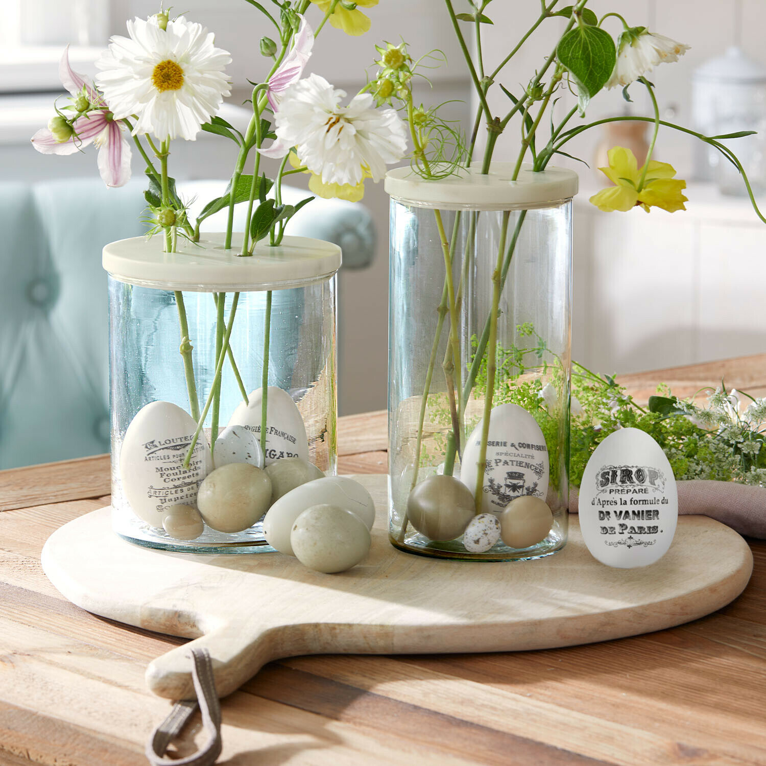 Glazen vazen met witte en beige decoratieve eieren en bloemen op een houten snijplank