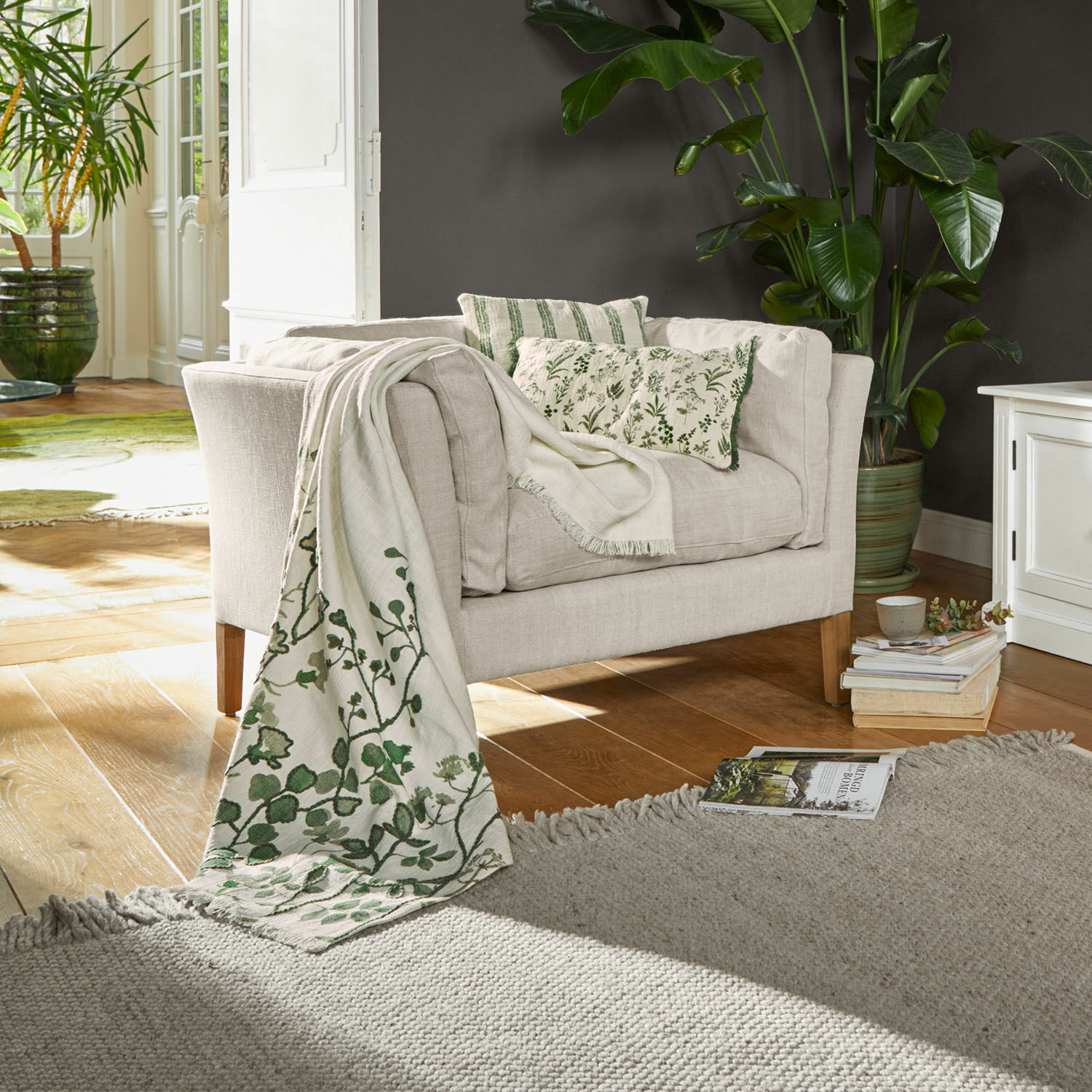 Beige stoffen bank met groene kussens en decoratief deken met plantenmotief in woonkamer