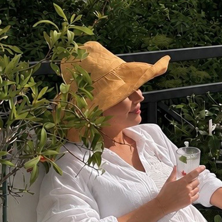 Vrouw in wit overhemd en gele strohoed ontspant op terras met drankje in hand
