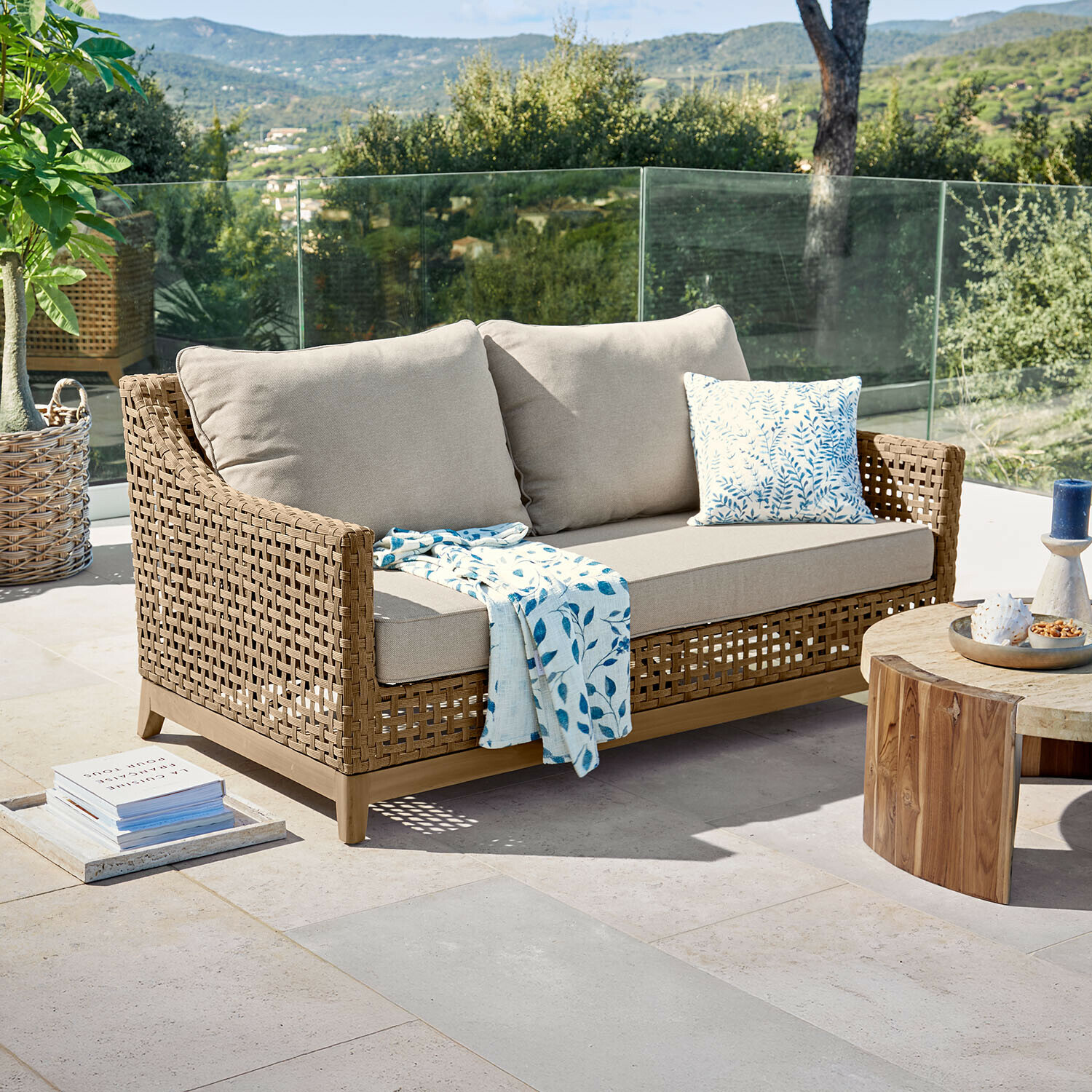 Buitensofa van natuurlijk gevlochten riet met beige kussens en blauw bladmotief deken