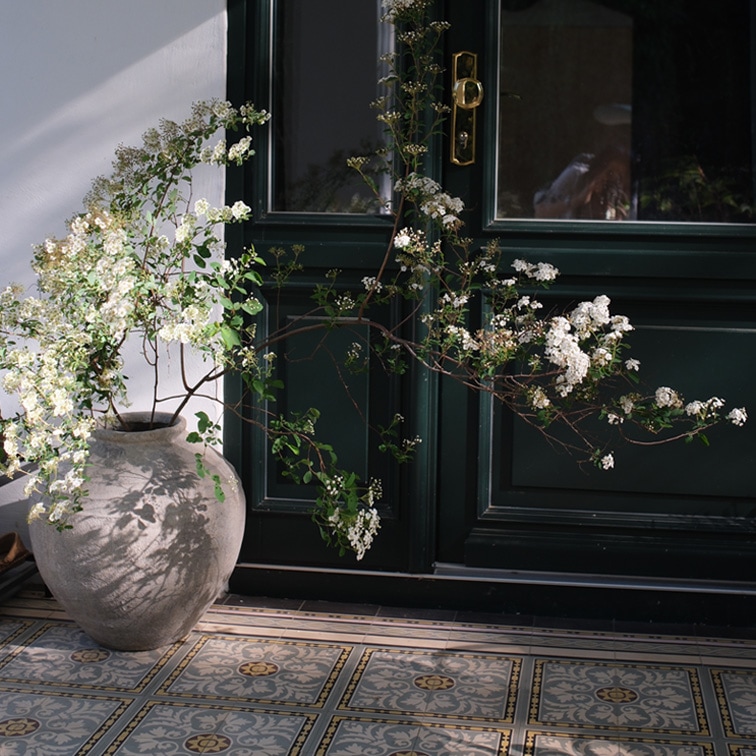 Grote keramische vaas met witte bloemen voor een donkere deur en decoratieve tegelvloer
