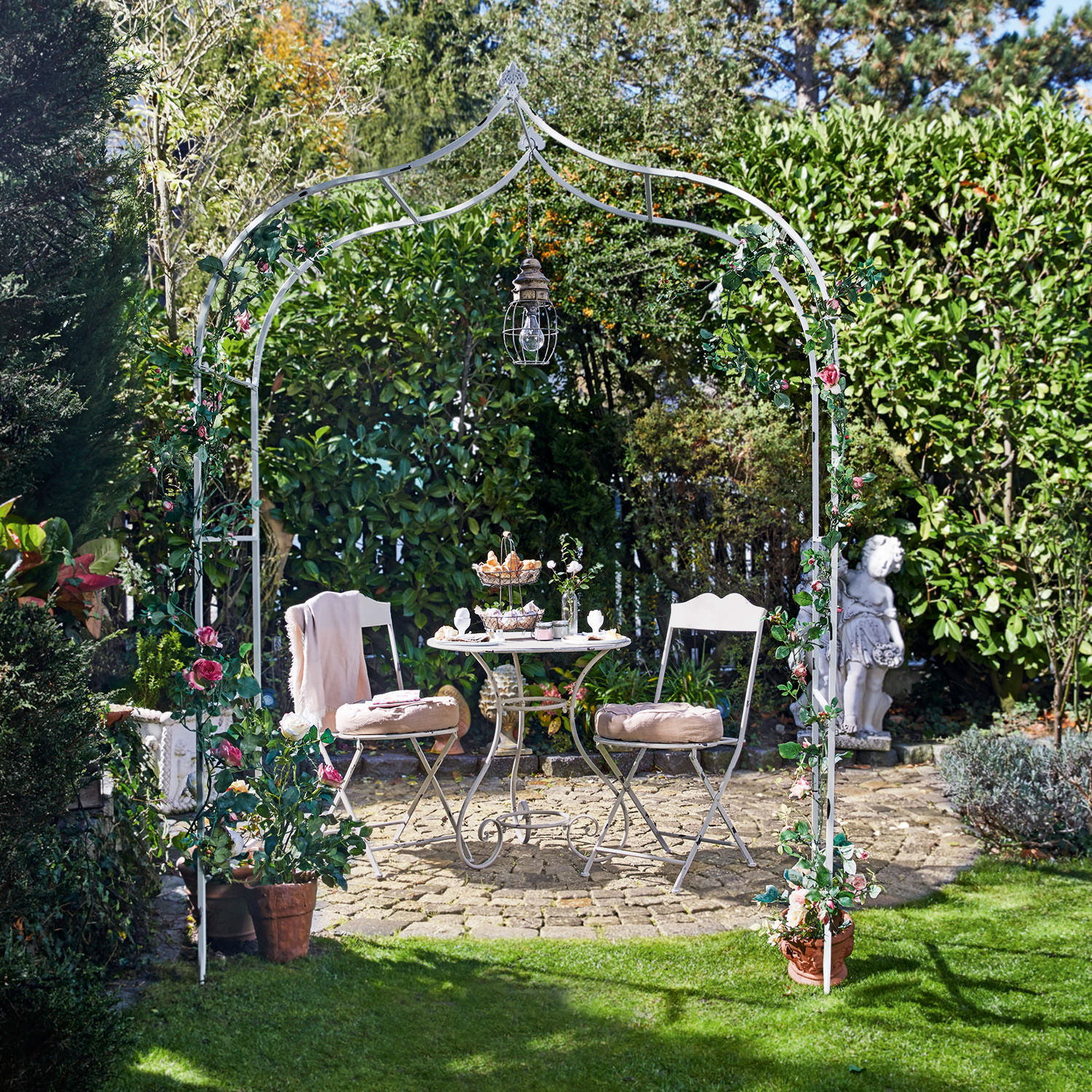 Rozenboog Dimarus Witte metalen tuinboog met roze bloemen en hanglamp, met tuinmeubilair op stenen terras