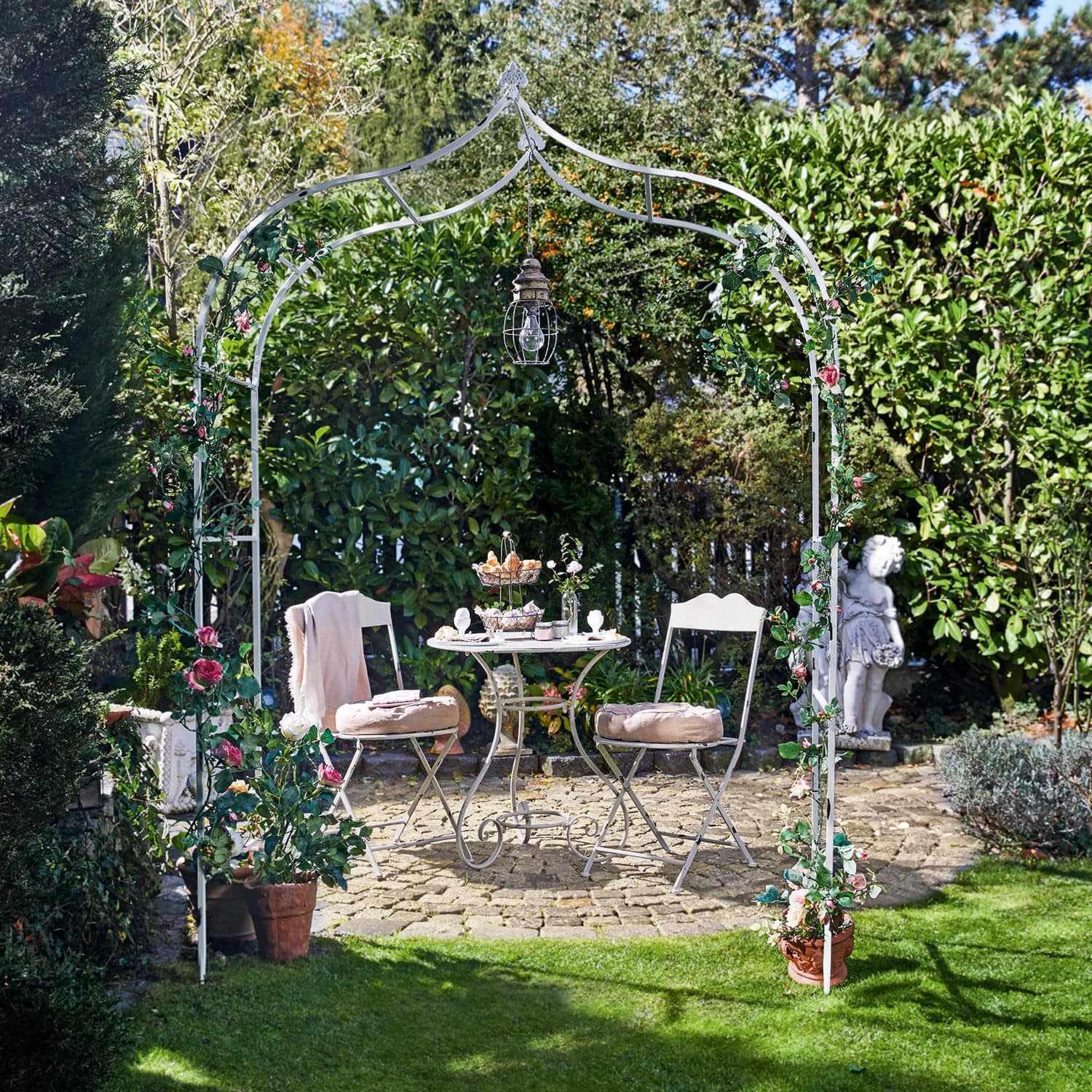 Rozenboog Dimarus Witte metalen tuinboog met roze bloemen en hanglamp, met tuinmeubilair op stenen terras