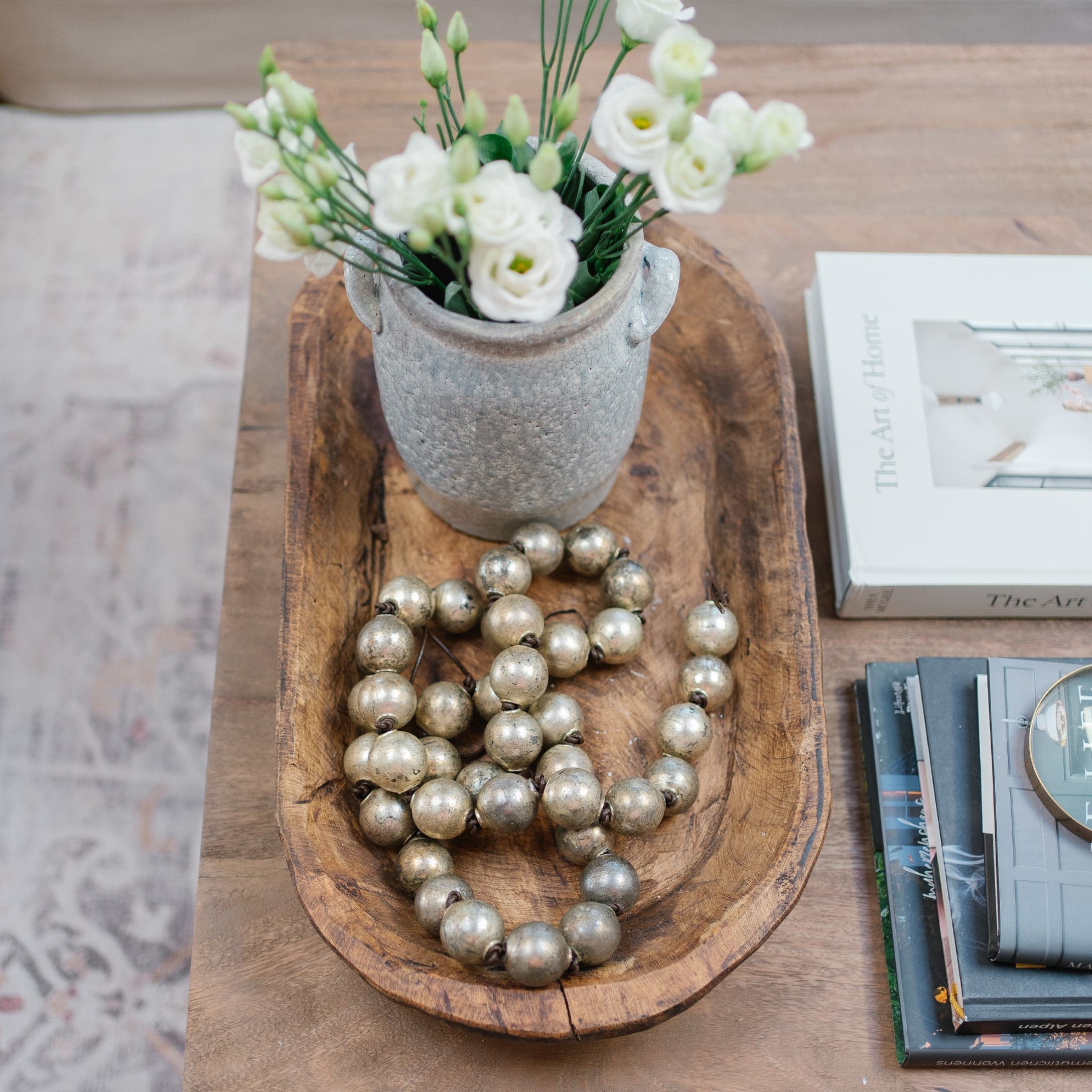 Houten schaal met grijze keramische vaas, witte bloemen en decoratieve metalen kralenketting op houten tafel