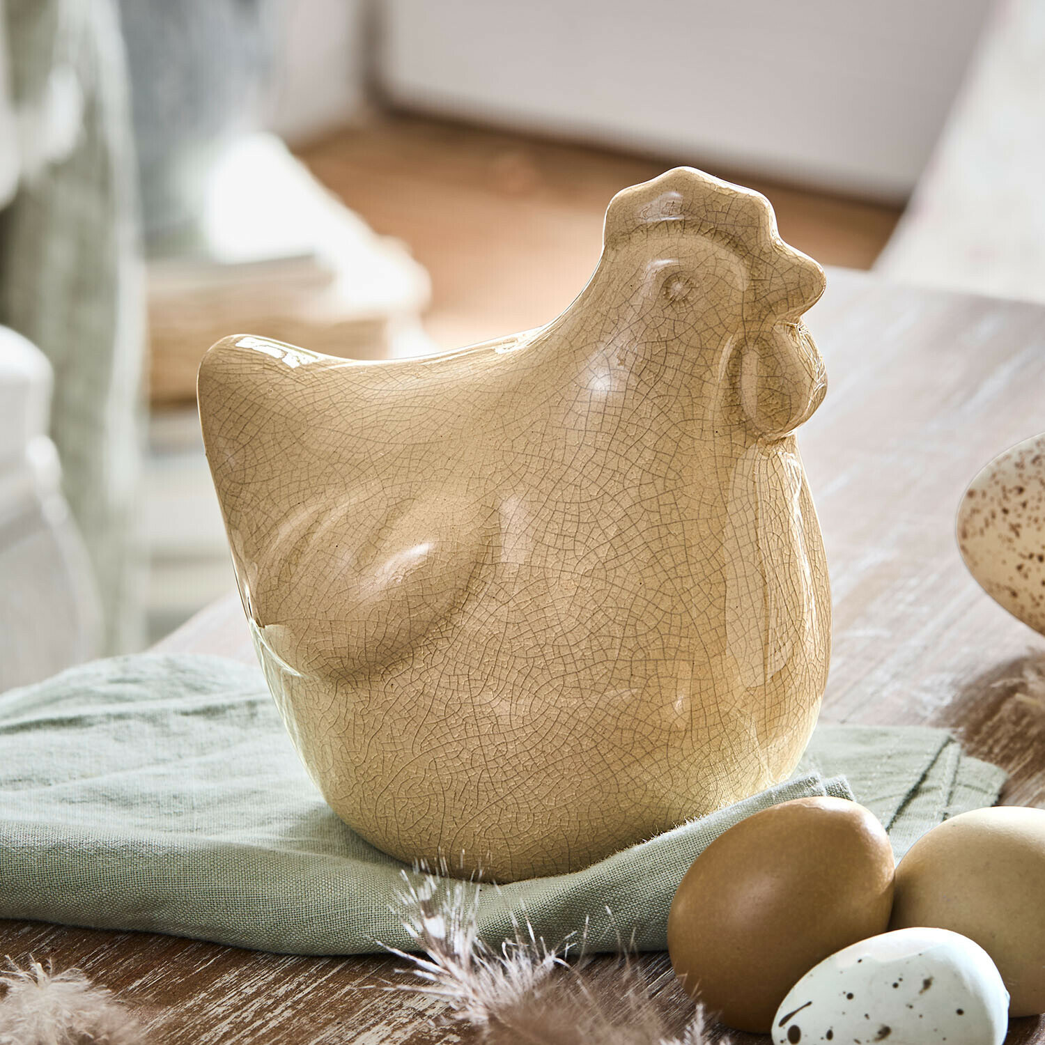 Decoratieve kip Roisenn Beige keramieken kippenfiguur met craquelé-effect op groene stof naast decoratieve eieren