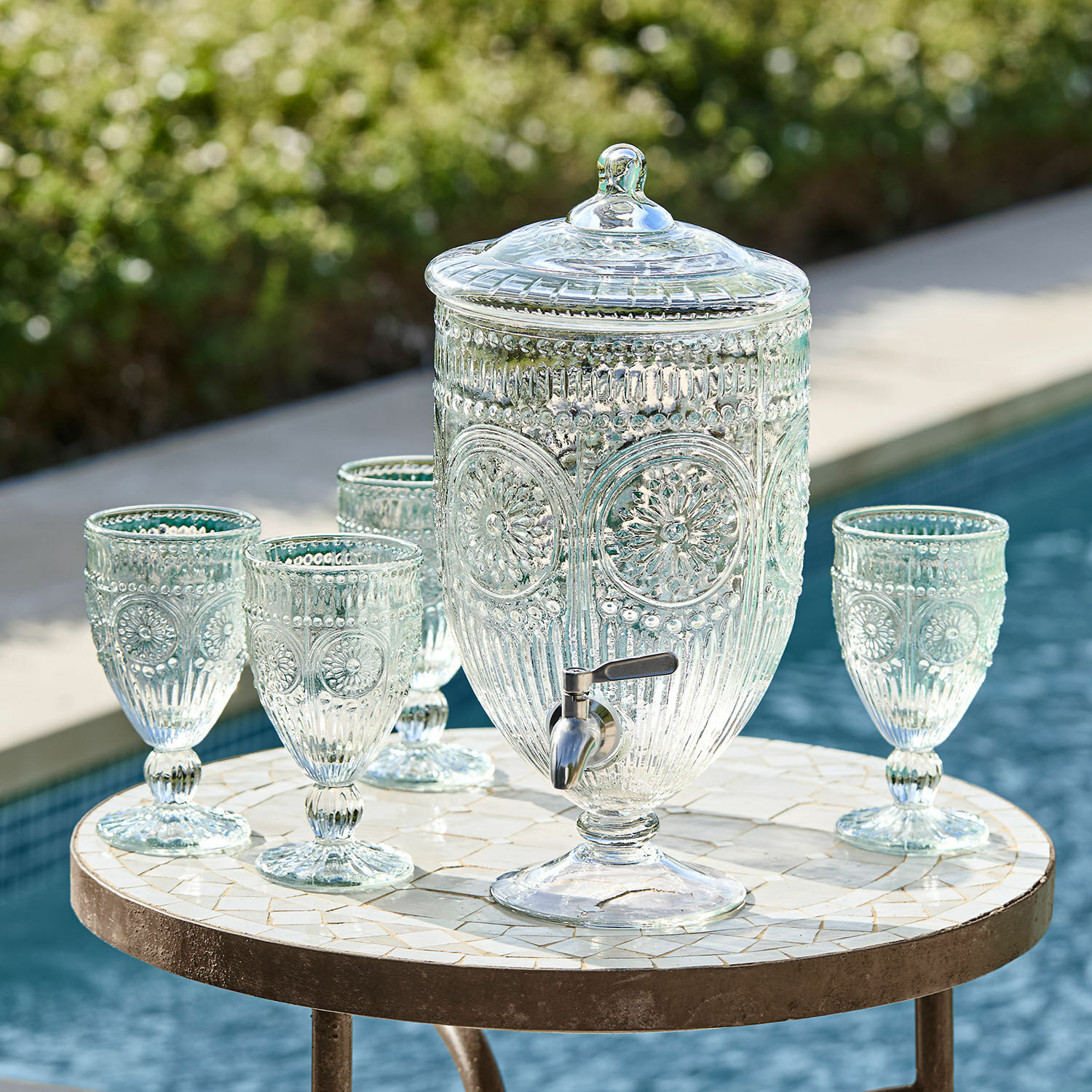 Glazen drankdispenser met deksel en kraan, met vier bijpassende glazen op een tafel bij het zwembad