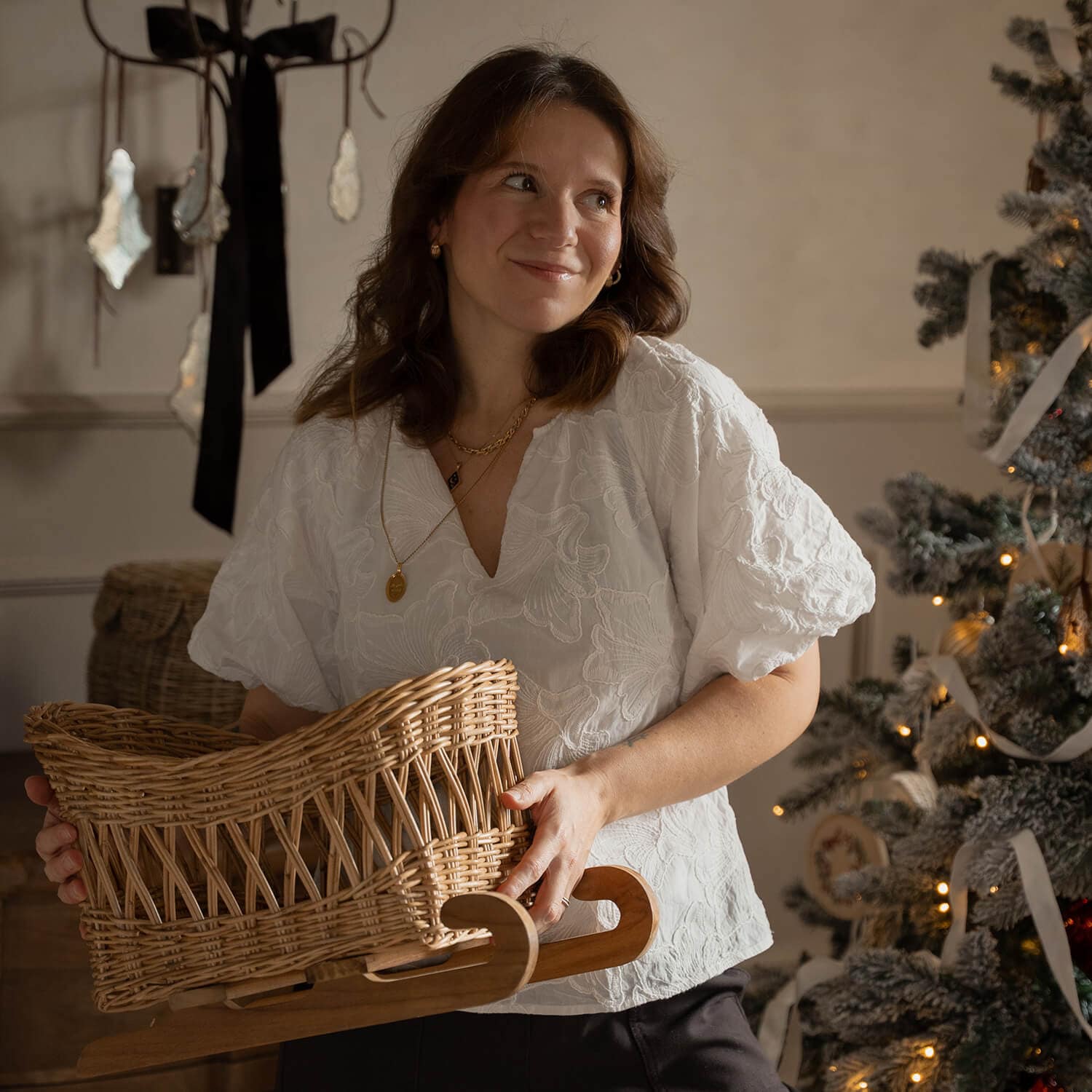 Vrouw in witte blouse houdt een rieten slee met houten onderstel vast bij een versierde kerstboom