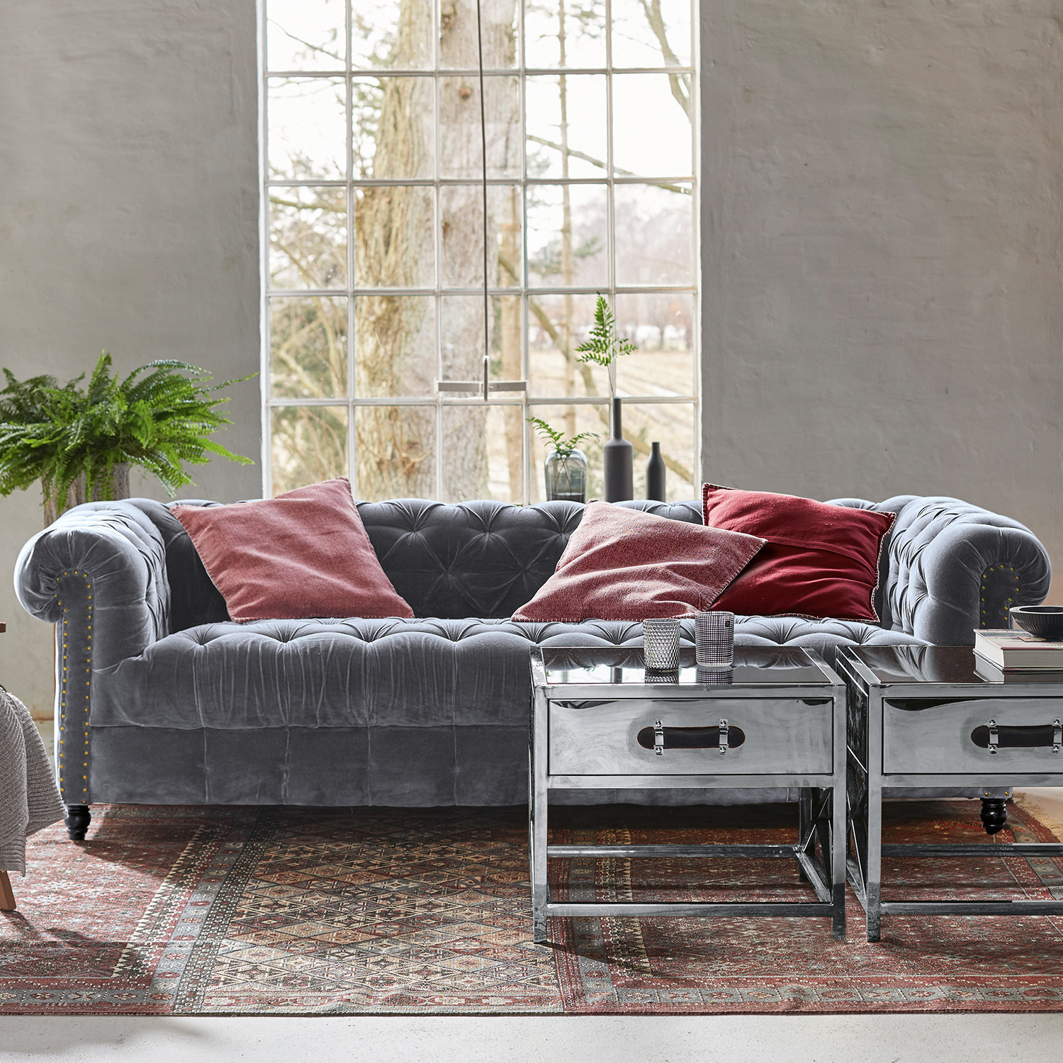 Bank Parpeville Grijze fluwelen Chesterfield bank met rode kussens en metalen salontafels op een decoratief vloerkleed