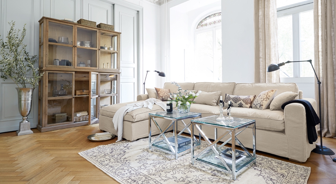 Gezellige woonkamer met beige bank, glazen tafels en houten vitrinekast op lichte vloer.