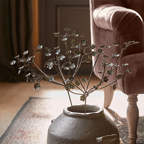 Decoratieve metalen tak met bloemen in grijze keramische vaas op tapijt naast roze fauteuil