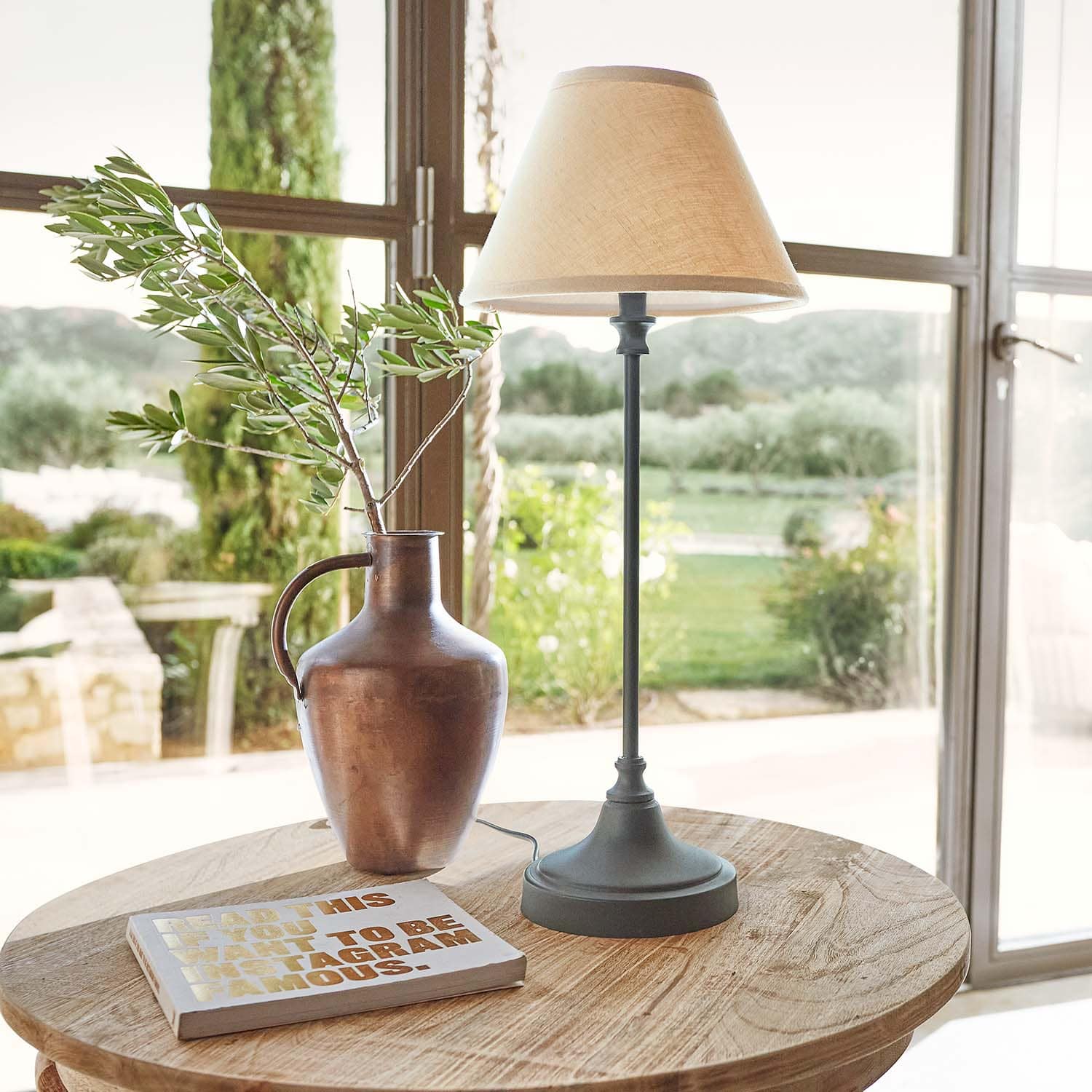 Metalen kan in koperkleur met tak en tafellamp met beige kap op houten tafel