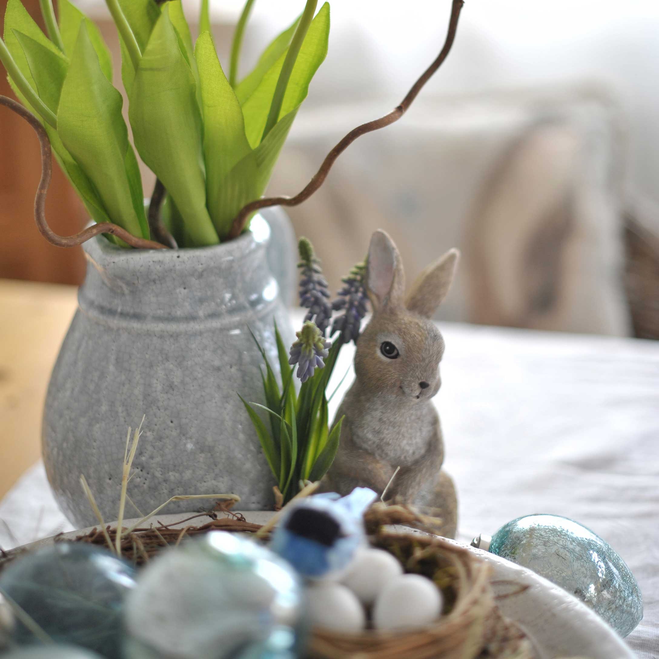 Grijze keramische vaas met groene bladeren en konijnenfiguur als paasdecoratie op tafel